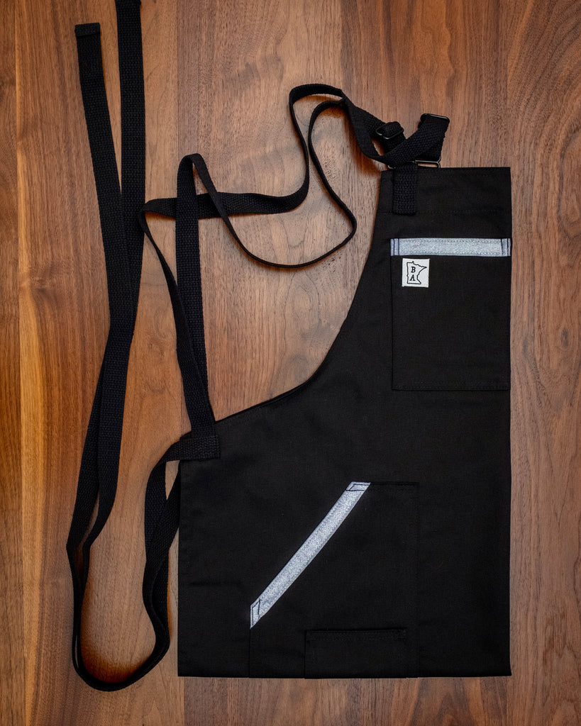Black cotton blended apron with metallic denim trim on the pockets and black strapping folded on a wooden surface.  The apron design 'Back to Battle' was manufactured in Minnesota by Craftmade Aprons.
