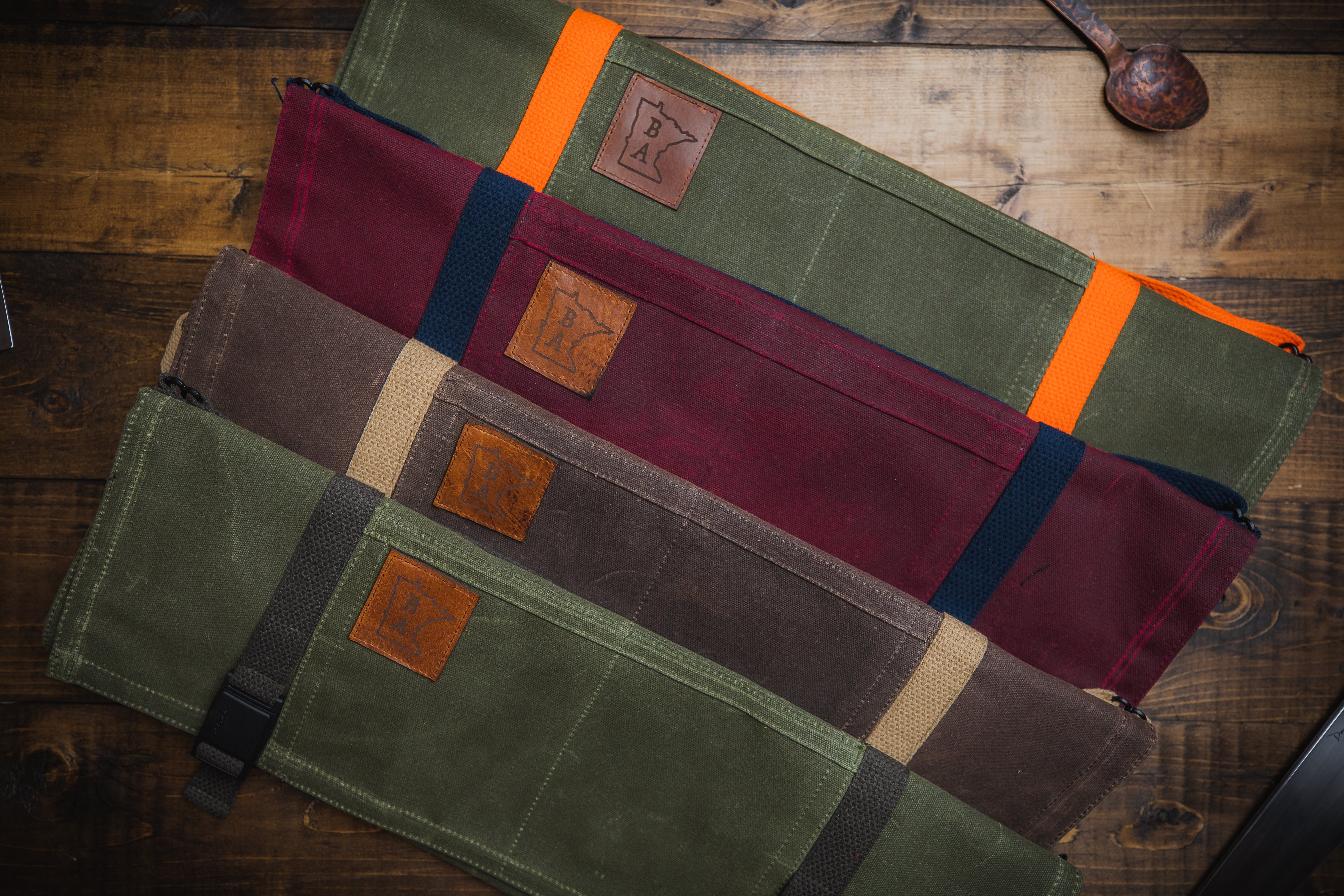 The Main Squeeze knife rolls displayed in colors of olive green with charcoal gray strapping, gray with natural cotton strapping, burgundy with navy blue strapping, and olive green with orange strapping. All are cotton knife rolls with an outer shell of tex -wax. All handcrafted in Minnesota by Craftmade Aprons.