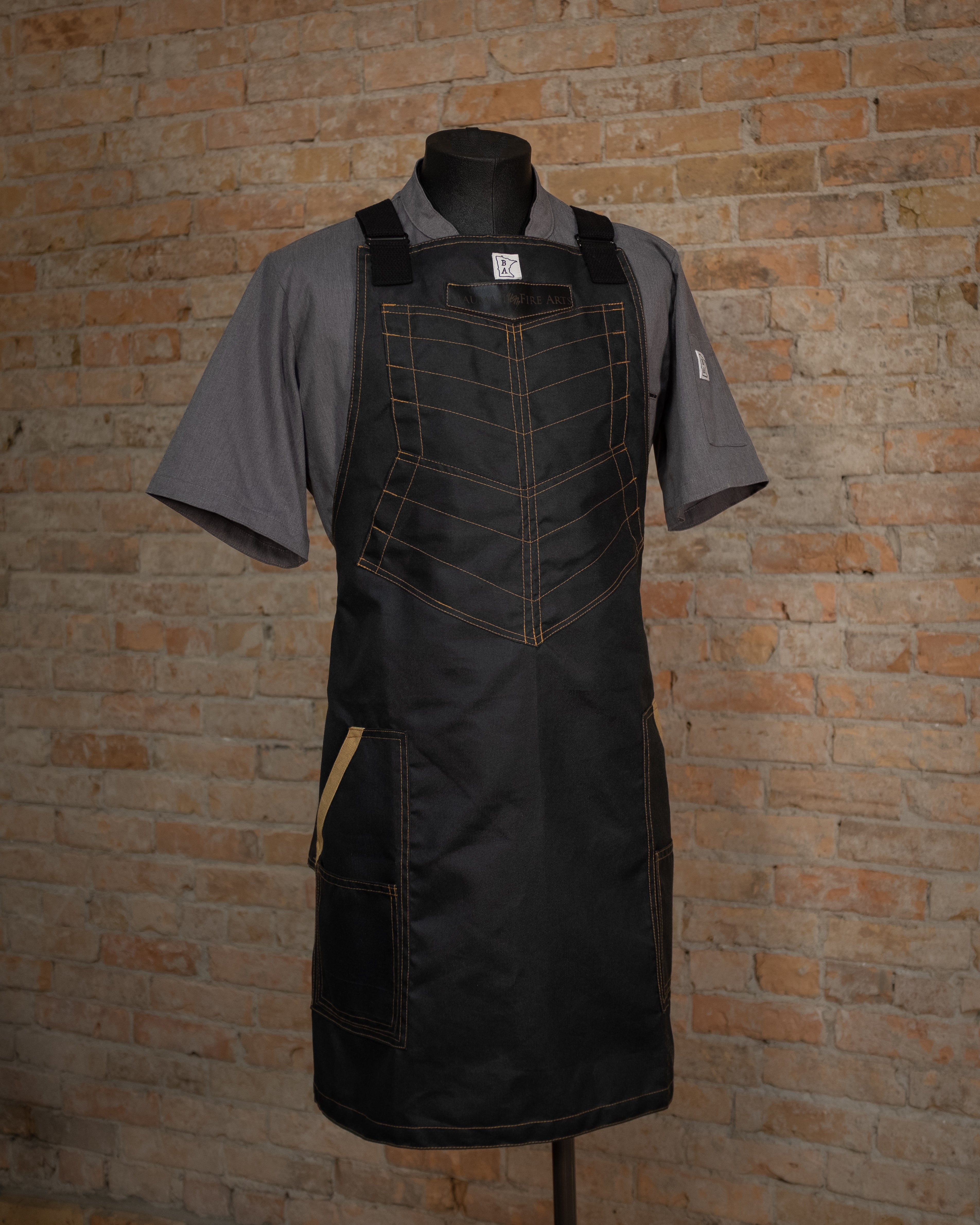 The heavy duty design 'BAMF', displayed on a mannequin over a gray chef  coat in front of a brick background. Both the chef coat and apron were designed and produced by Craftmade Aprons, Minnesota.
