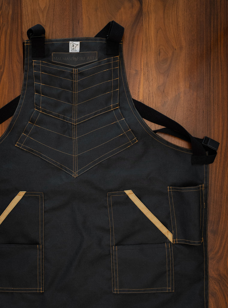 Product shot of a heavy-duty, protective Kevlar apron in black with yellow threading on a wooden surface. The BAMF is the perfect apron for knife makers. Made in collaboration between Craftmade Aprons, MN, and Maumasi Fire Arts, WA.