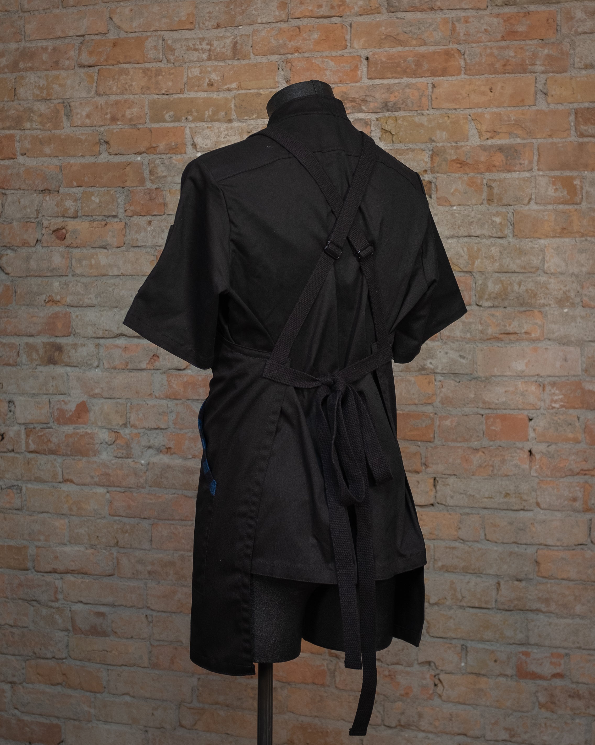 Cotton crossback apron from Craftmade Aprons, Minnesota, displayed on a mannequin over a black chef coat. The apron Black and Blue II is shown from the back, showing the black crossback strapping.