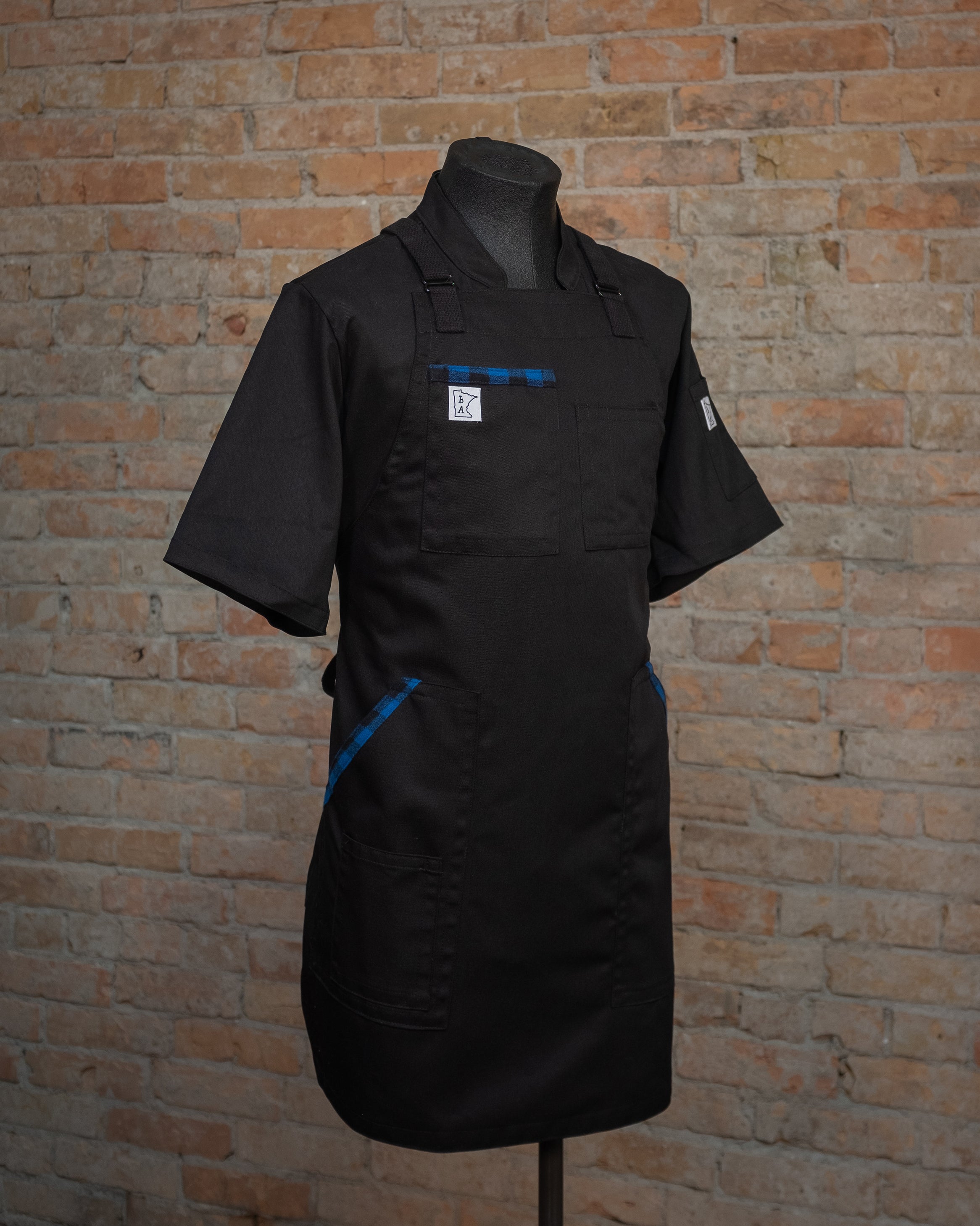 The cotton blended apron design Black and Blue II displayed on a mannequin over a black chef coat.  The cotton blend gives the second apron of the Project Black and Blue collection a slight stretch. Both the chef coat and the apron were designed and produced by Craftmade Aprons located in Minnesota. 