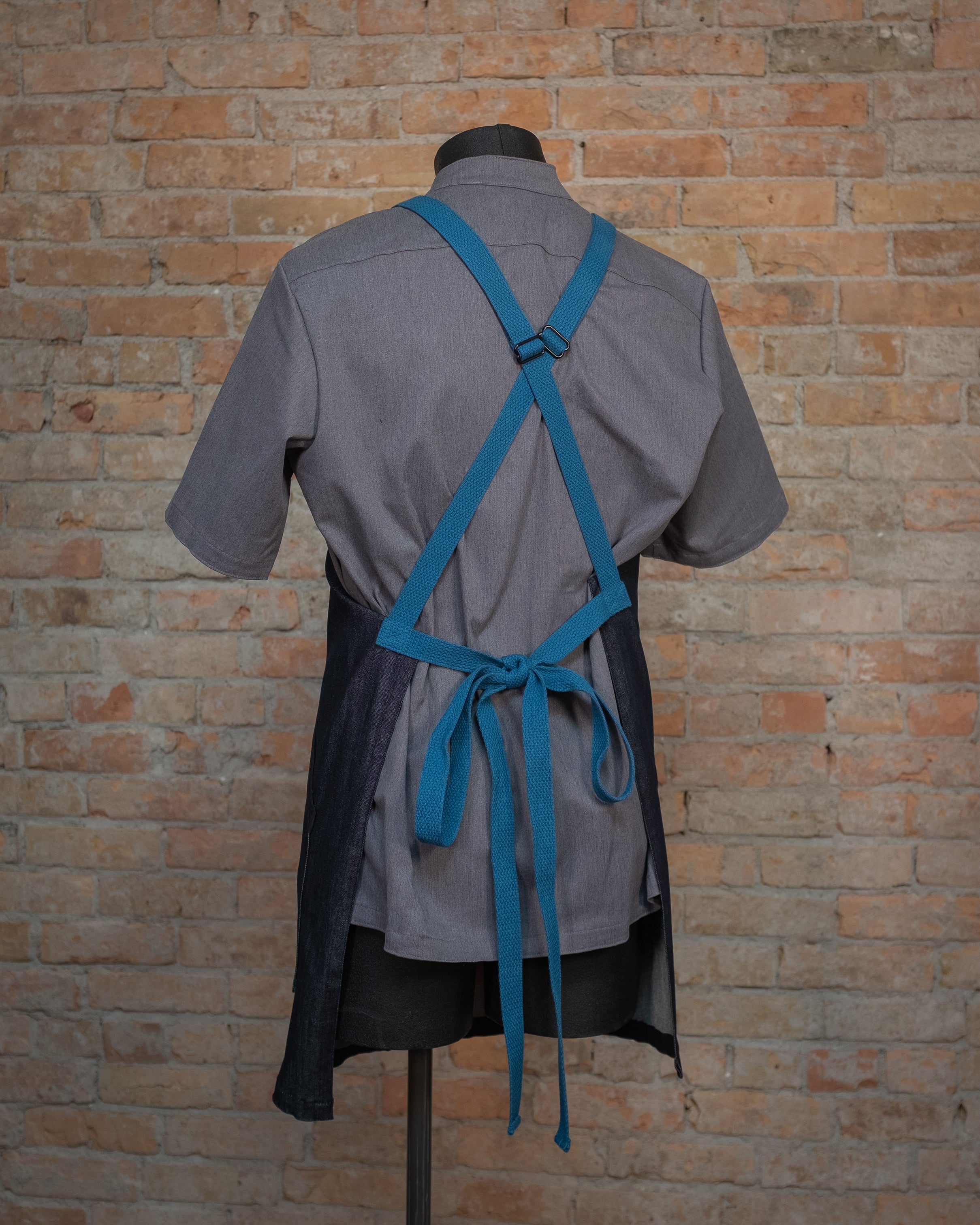 Denim crossback apron from Craftmade Aprons, Minnesota, displayed on a mannequin over a gray chef coat. The apron Black & Blue Denim is shown from the back, revealing the signature blue crossback strapping.