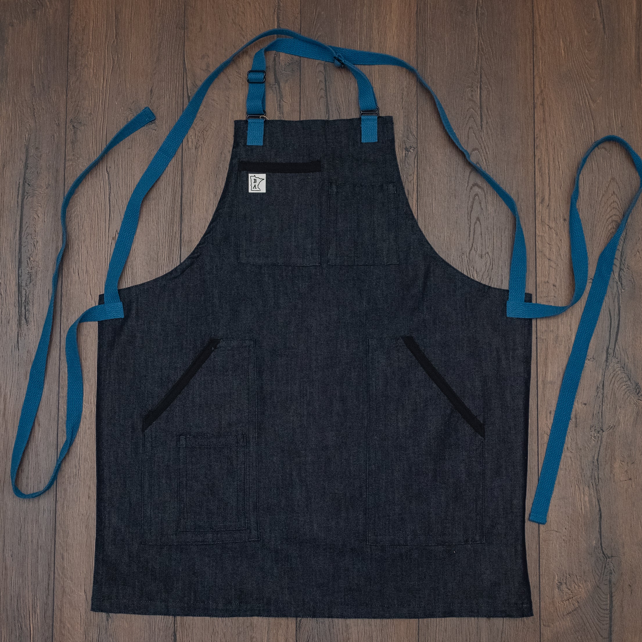 The denim apron design of the Black and Blue collection laid out flat on a wooden surface, making visible the blue strapping and black lined pockets. Black and Blue Denim was designed and sold by Craftmade Aprons, based in Minnesota. Sold in sizes of standard, Large, and XL.