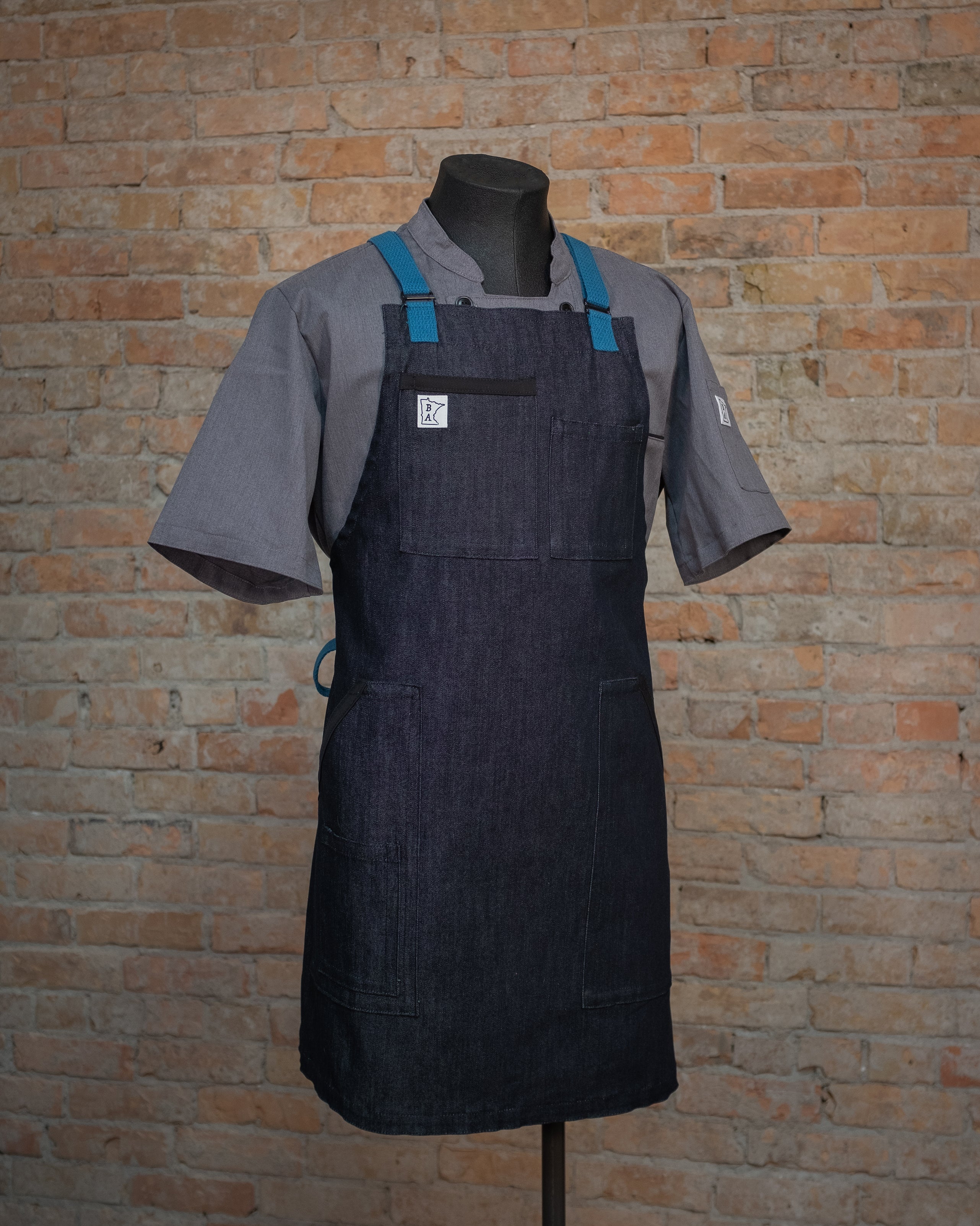 The denim apron 'Black and Blue', displayed on a mannequin over a gray chef coat. Both the chef coat and apron were designed and produced by Craftmade Aprons in Minnesota. This  apron helps represent Project Black and Blue, with 50% of revenue going directly to people in the service industry. 