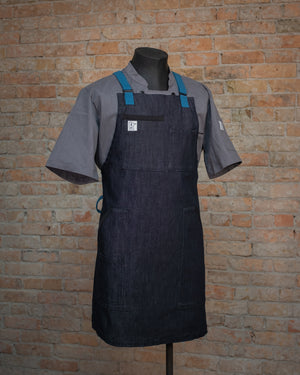 The denim apron 'Black and Blue', displayed on a mannequin over a gray chef coat. Both the chef coat and apron were designed and produced by Craftmade Aprons in Minnesota. This  apron helps represent Project Black and Blue, with 50% of revenue going directly to people in the service industry. 