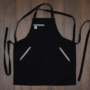 Black cotton blend apron with metallic denim details on the pockets and black strapping laid out flat on a wooden surface. Design 'Back to Battle' created by Cratfmade Aprons, based in Minnesota.