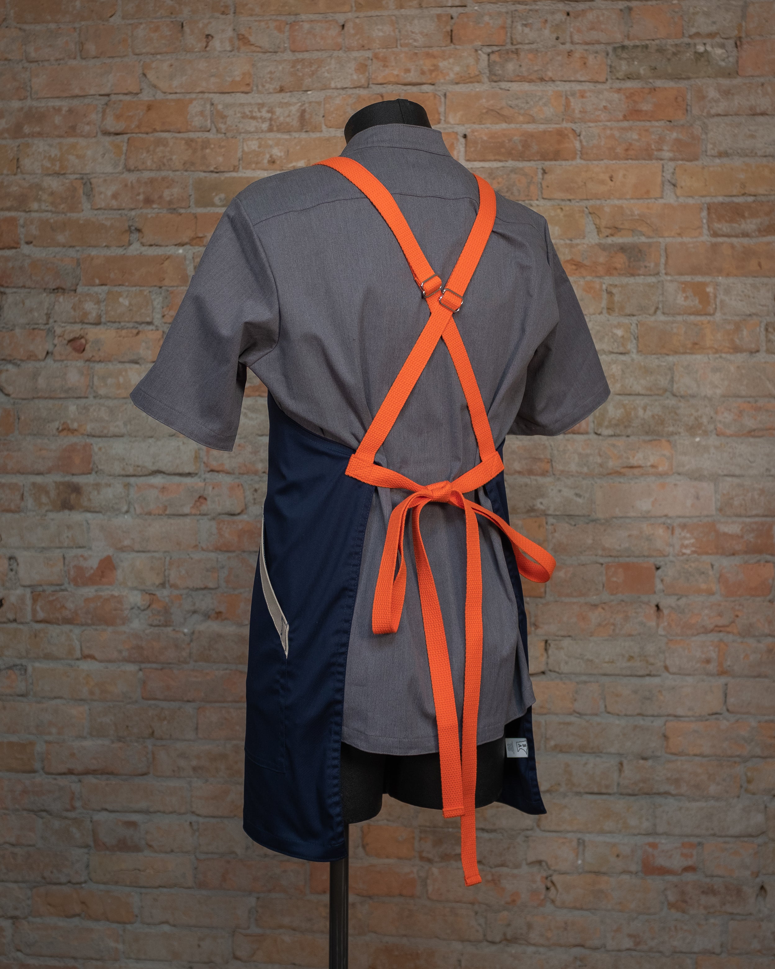 Blended fabric crossback apron from Craftmade Aprons, Minnesota, displayed on a mannequin over a gray chef coat. The apron 'Bert' is shown from the back, showing the orange crossback strapping in detail.