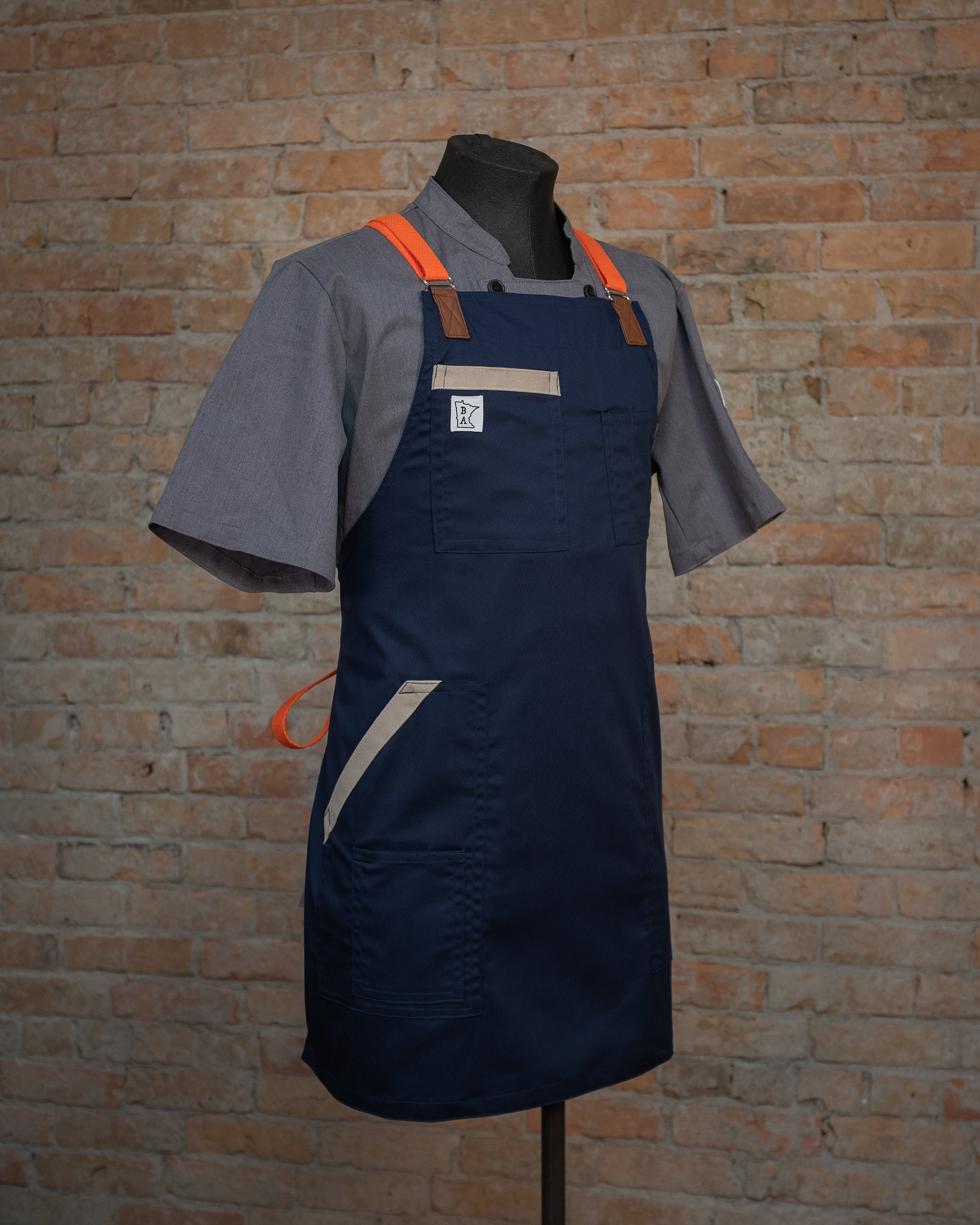 The blended fabric apron design 'Bert', displayed on a mannequin over a gray chef coat. Both the chef coat and apron were designed and produced by Craftmade Aprons in Minnesota.