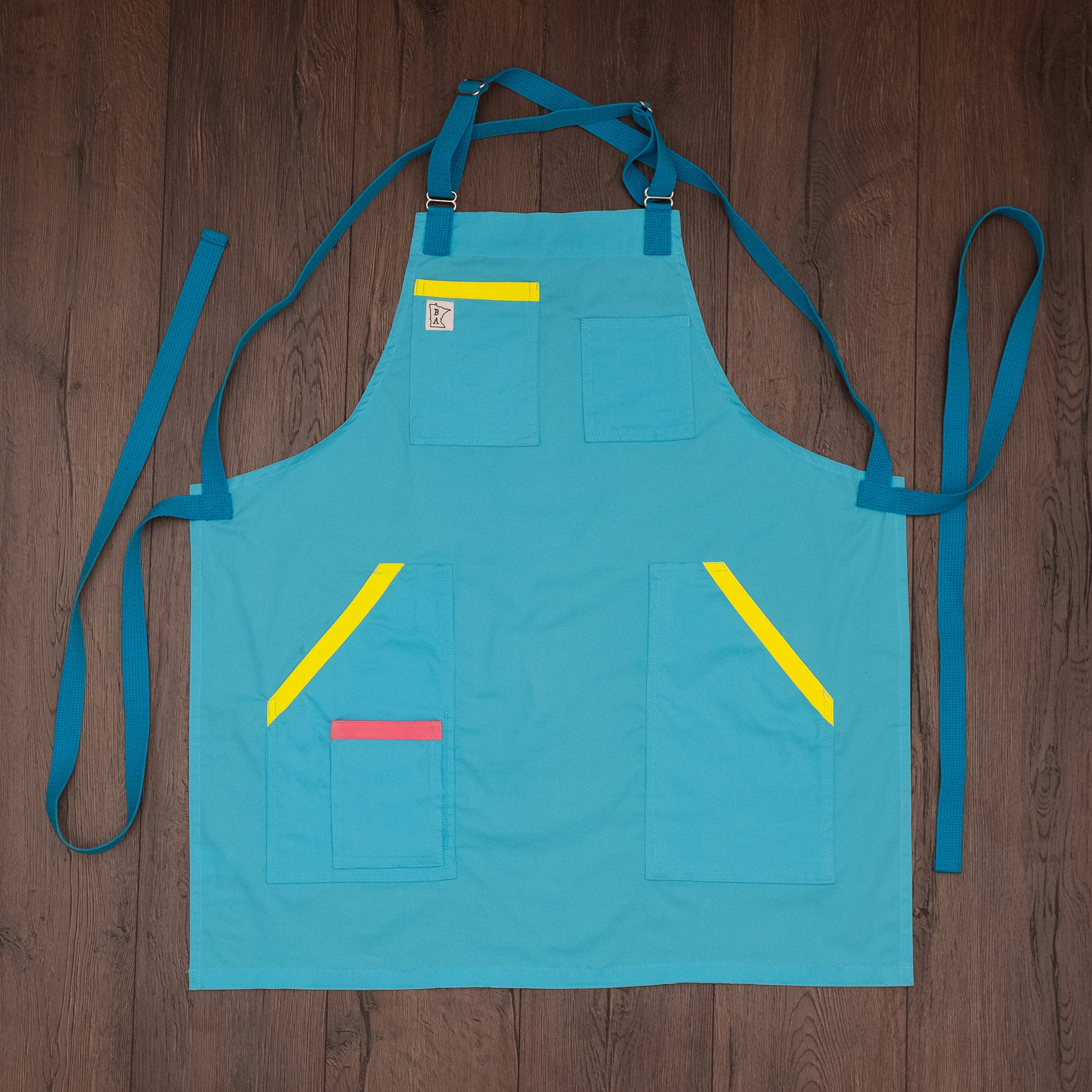 Ocean blue  Italian cotton apron with bright yellow and pink trimmed pockets and blue strapping laid out flat on a wooden surface. Design 'Billy Ocean' by Craftmade Aprons, based in Minnesota. 