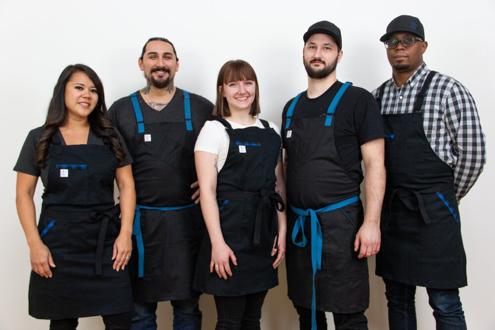 Five individuals wearing the "Black and Blue' apron, showing off the black base and signature blue strapping. 