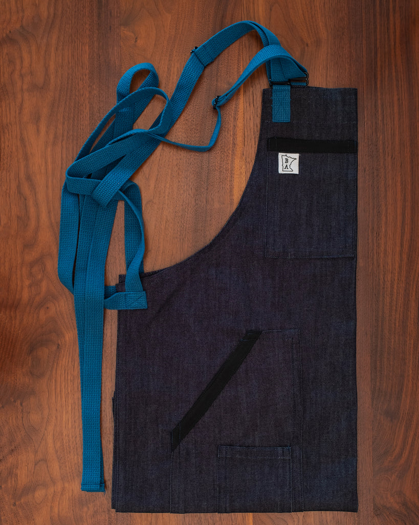 Project Black and Blue denim apron with signature blue strapping folded on a wooden surface. The apron design "Black and Blue Denim' was manufactured in Minnesota by Craftmade Aprons. 50% of revenue goes directly to those in the service industry.
