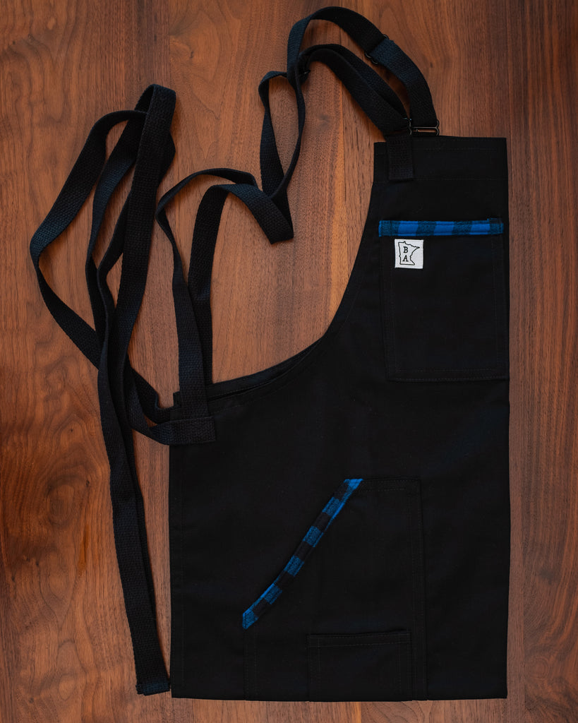 Cotton blend apron with blue flannel accents on the pockets with black strapping folded on a wooden surface. The apron design Black and Blue II was manufactured in Minnesota by Craftmade Aprons. 50% of all revenue goes directly to the Project Black and Blue fund.