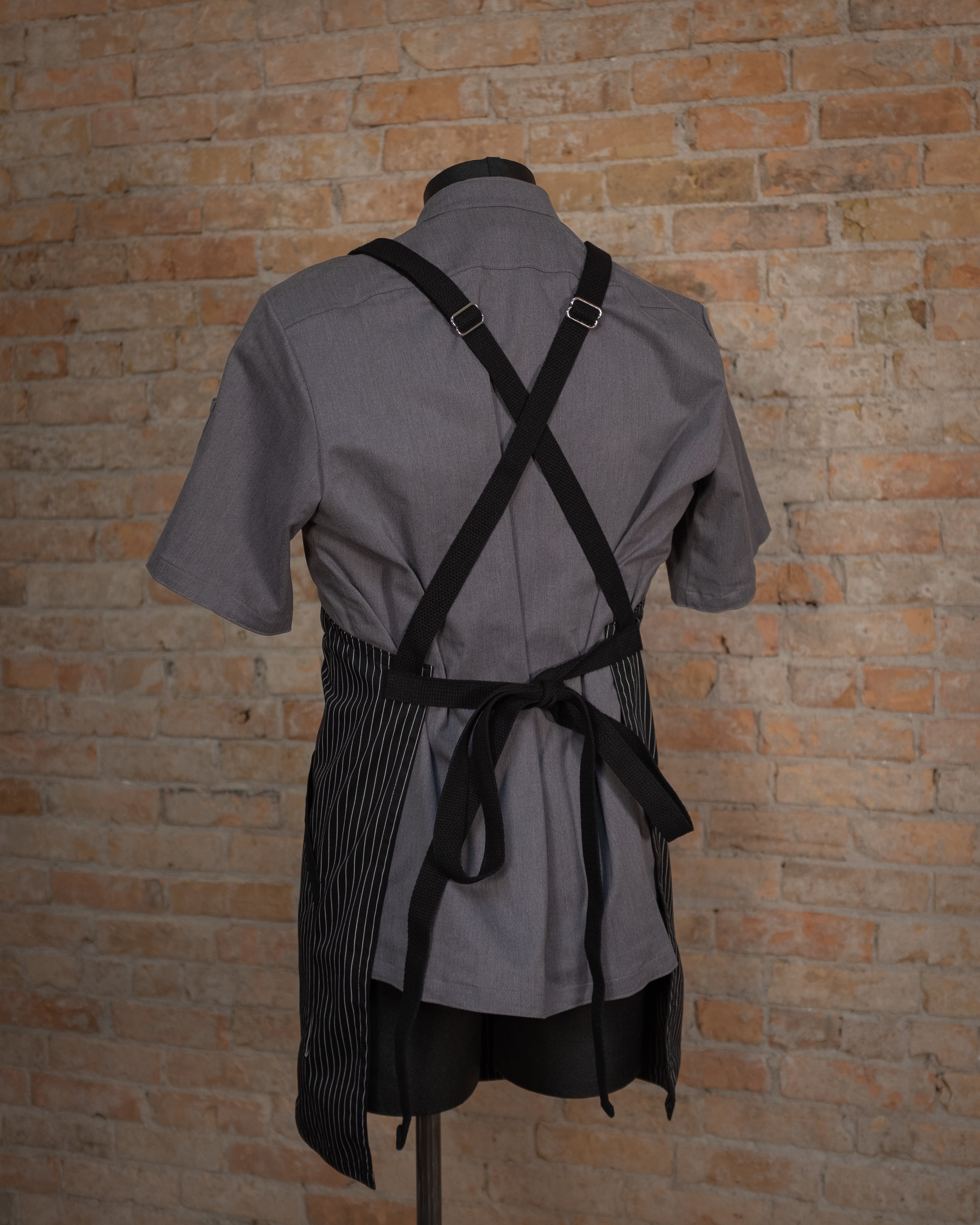 Poly/cotton twill crossback apron from Craftmade Aprons, Minnesota, displayed on a mannequin over a gray chef coat. The apron Culinary Gangster is shown in black from the back, displaying the black crossback strapping.