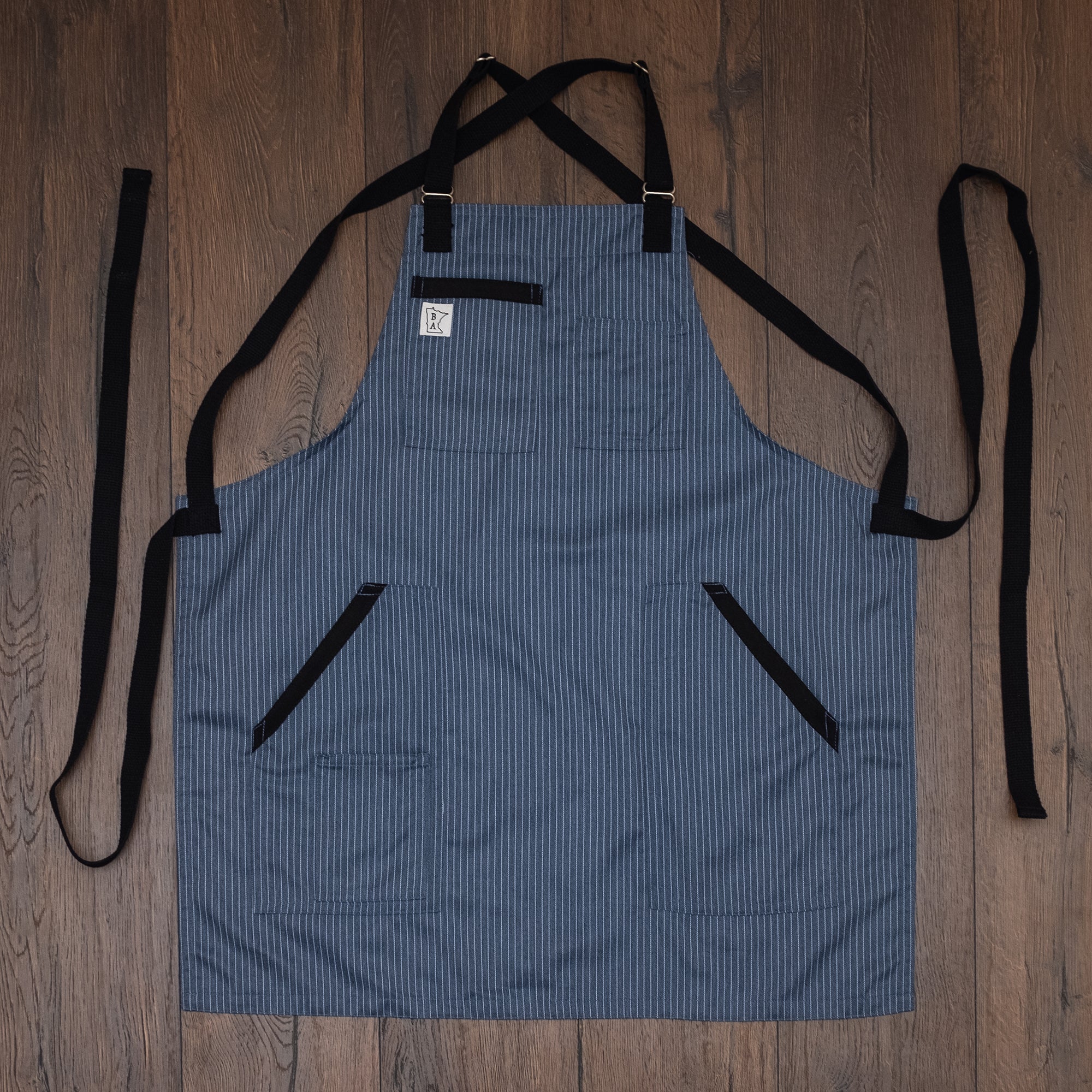 The poly/cotton twill apron design Culinary Gangster with black accents on the pockets and black strapping laid out flat on a wooden surface. Produced by Craftmade Aprons, based in Minnesota.