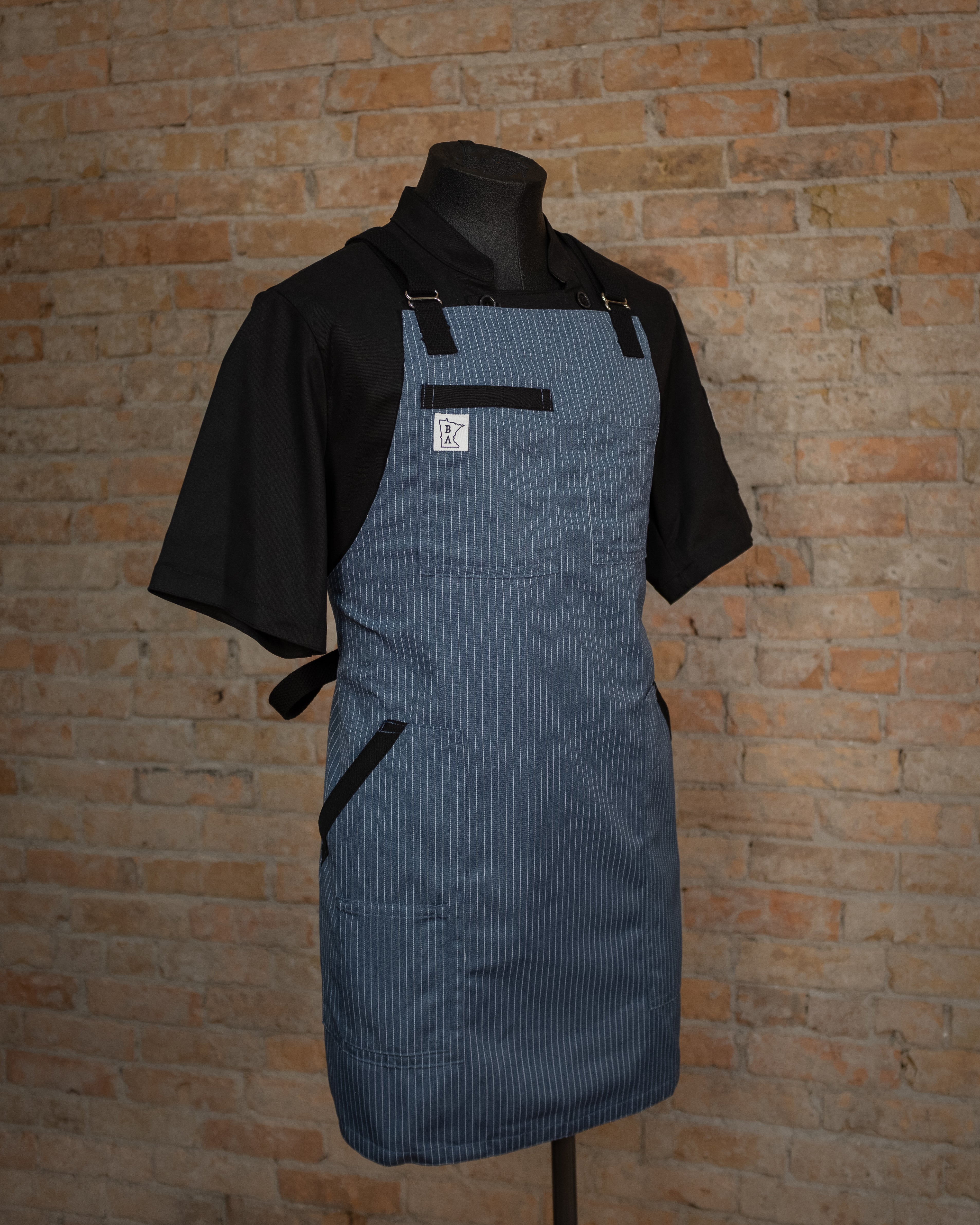 Poly/cotton twill apron with a blue base and white pinstripes displayed on a mannequin over a black chef coat. Both the chef coat and Culinary Gangster apron were designed by Craftmade Aprons, Minnesota. 