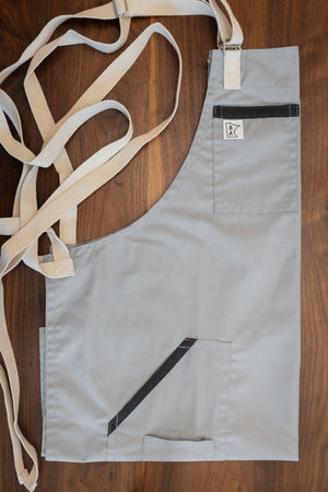 Cotton/poly blend apron with a cool gray base, charcoal gray trim, and natural colored strapping folded on a wooden surface. The apron design Cloudy With a Chance was manufactured in Minnesota by Craftmade Aprons. 