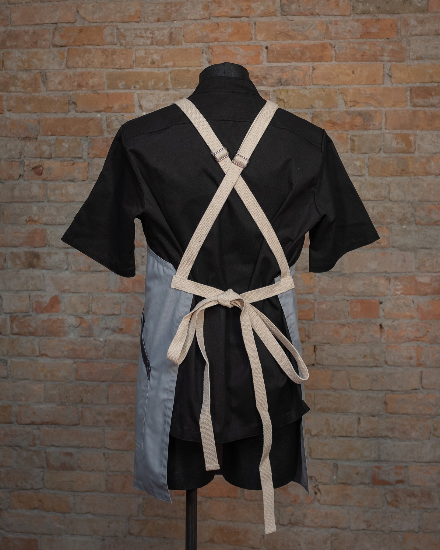Lightweight cotton/poly crossback apron from Craftmade Aprons, Minnesota, displayed on a mannequin over a black chef coat. The apron Cloudy With a Chance is shown from the back, displaying the natural colored crossback strapping.