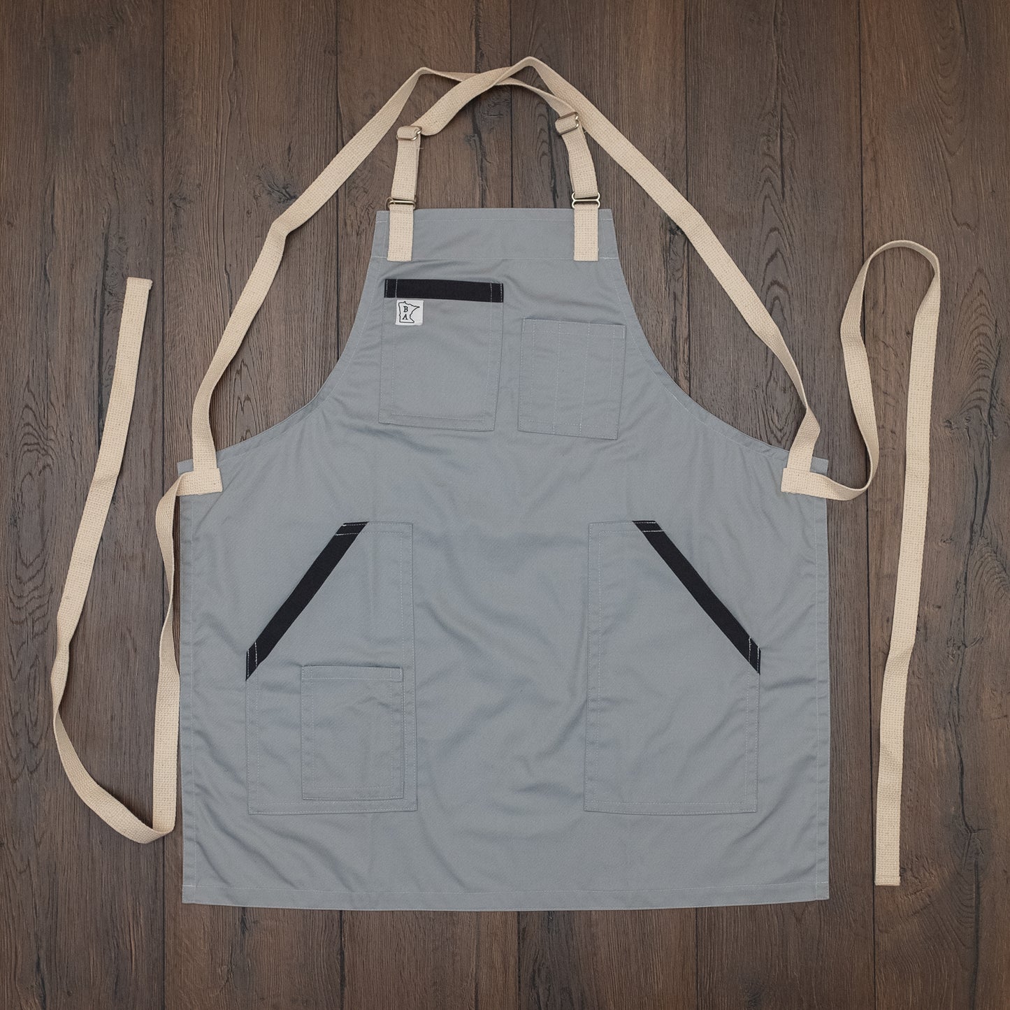 Cotton/poly apron with charcoal trim on the pockets and natural colored strapping laid out flat on a wooden surface. Design Cloudy With a Chance by Craftmade Aprons, based in Minnesota.