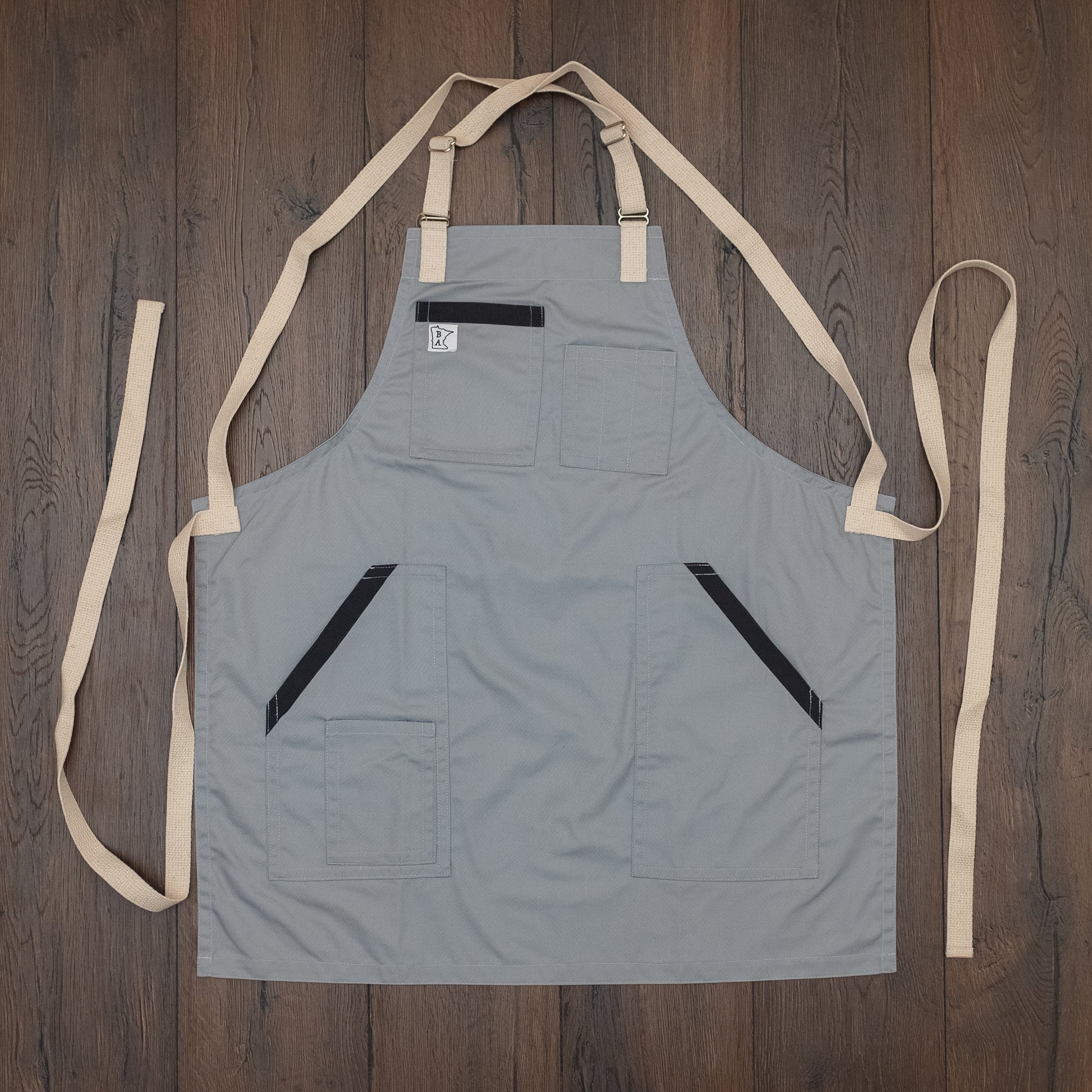 Cotton/poly apron with charcoal trim on the pockets and natural colored strapping laid out flat on a wooden surface. Design Cloudy With a Chance by Craftmade Aprons, based in Minnesota.