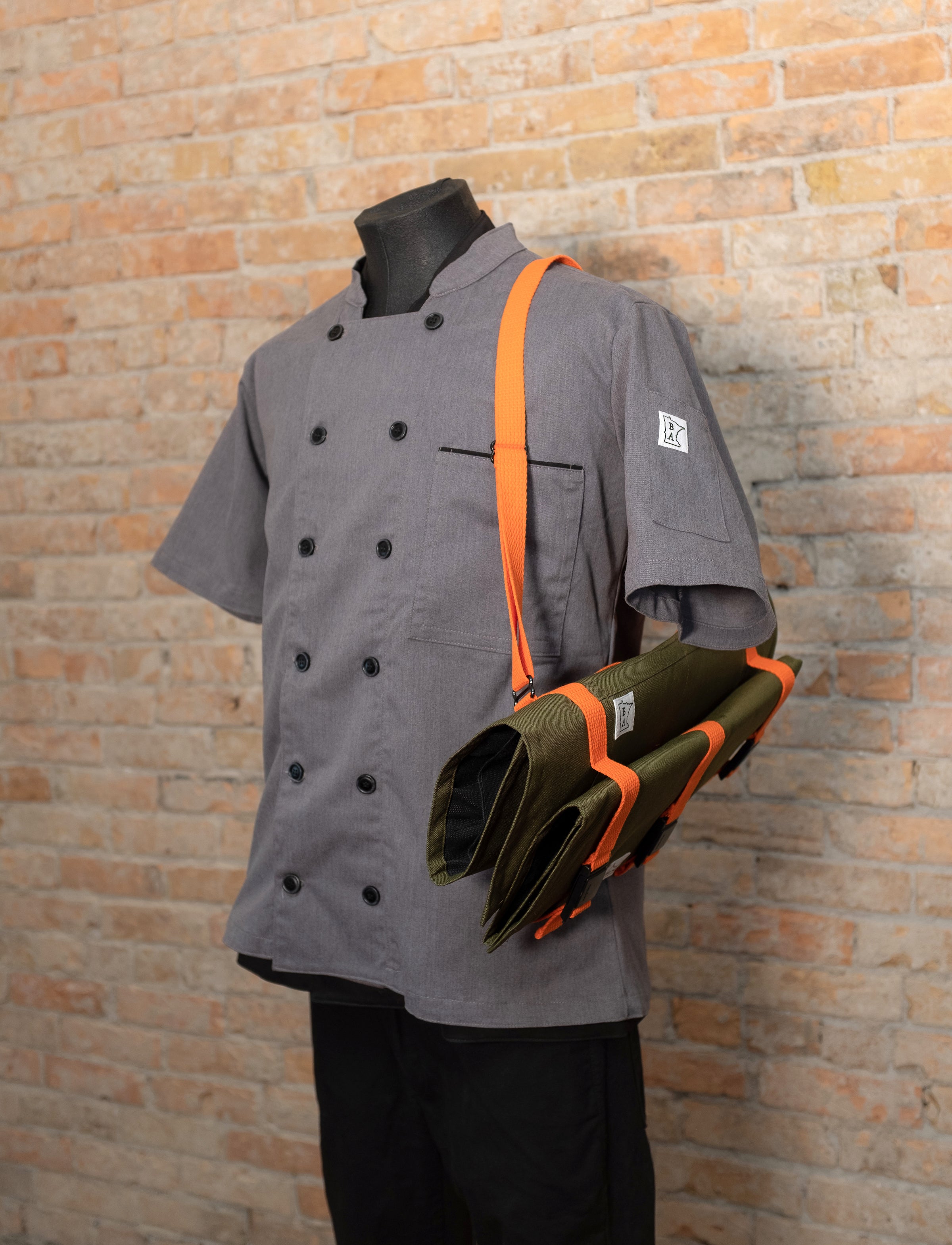 The Ottertex Edition Olive/Orange Main Squeeze and Side Hustle knife roll buckled together and slung over the shoulder of a mannequin wearing a gray chef coat. Both of the knife rolls and the chef coat were produced in Minnesota by Craftmade Aprons.