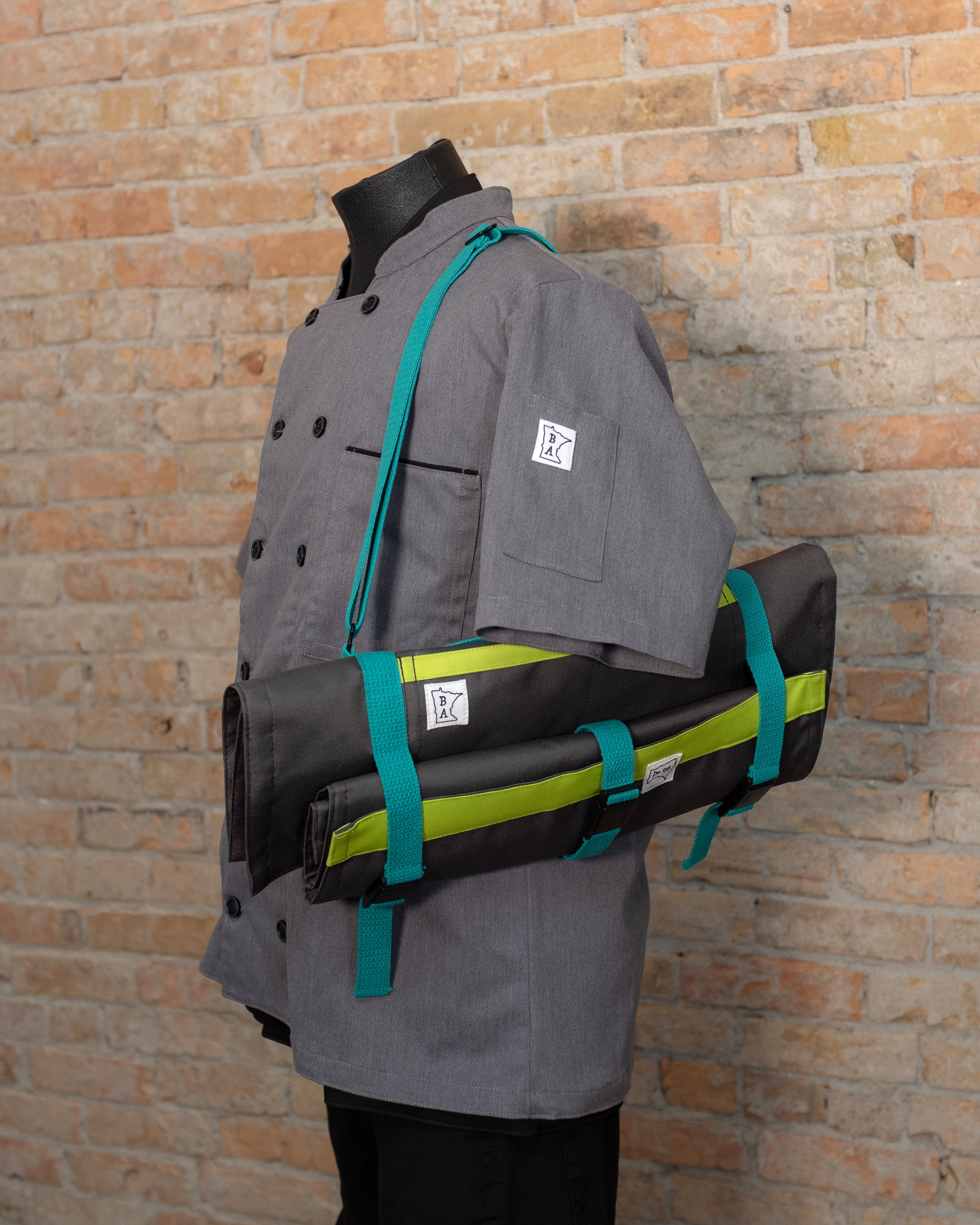 Main Squeeze Ottertex Edition /Totally Rad knife roll with matching Side Hustle knife roll slung over the shoulder of a mannequin in a gray chef coat. All items were designed by Craftmade Aprons, based in Minnesota.