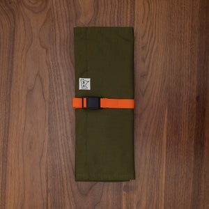 Cotton olive green  knife roll protected in tex-wax folded with the bright orange strapping on a wooden surface. The knife roll design Side Hustle was manufactured in Minnesota by Craftmade Aprons.