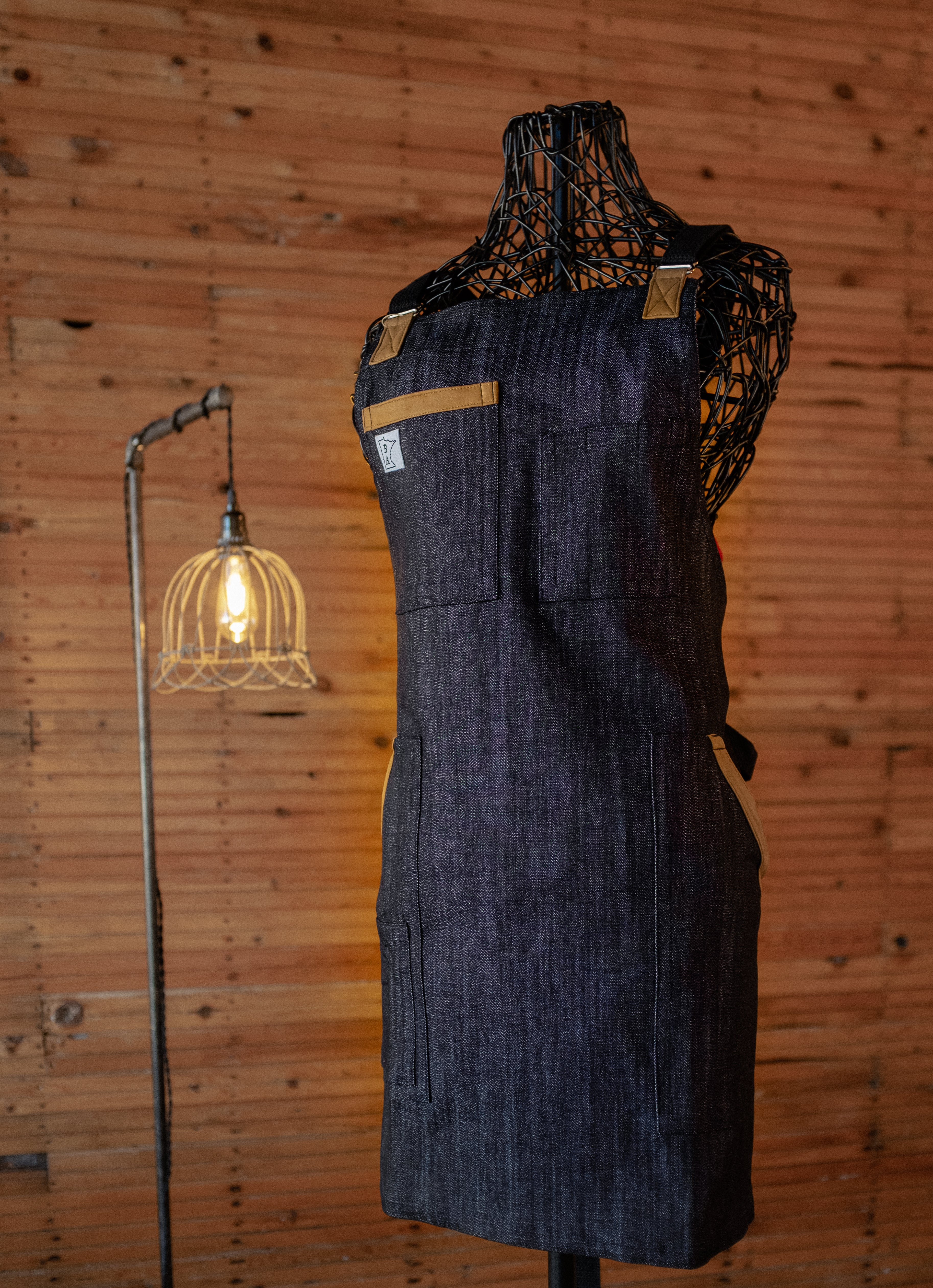 The denim apron design Denim and Deerhide displayed on a mannequin in front of a wood background. The apron was designed and produced by Craftmade Aprons, Minnesota.