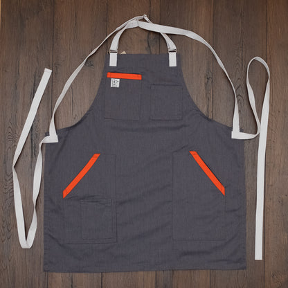 The cotton apron design Dreamsicle with a gray base and vibrant orange trimming on the pockets laid out flat on a wooden surface. Design Dreamsicle by Craftmade Aprons, based in Minnesota.