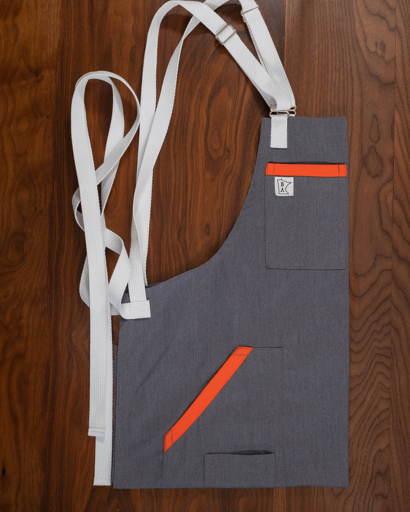 Cotton apron with a gray base, orange accents on the pockets, and bright white strapping folded on a wooden surface. The apron design Dreamsicle was manufactured in Minnesota by Craftmade Aprons.