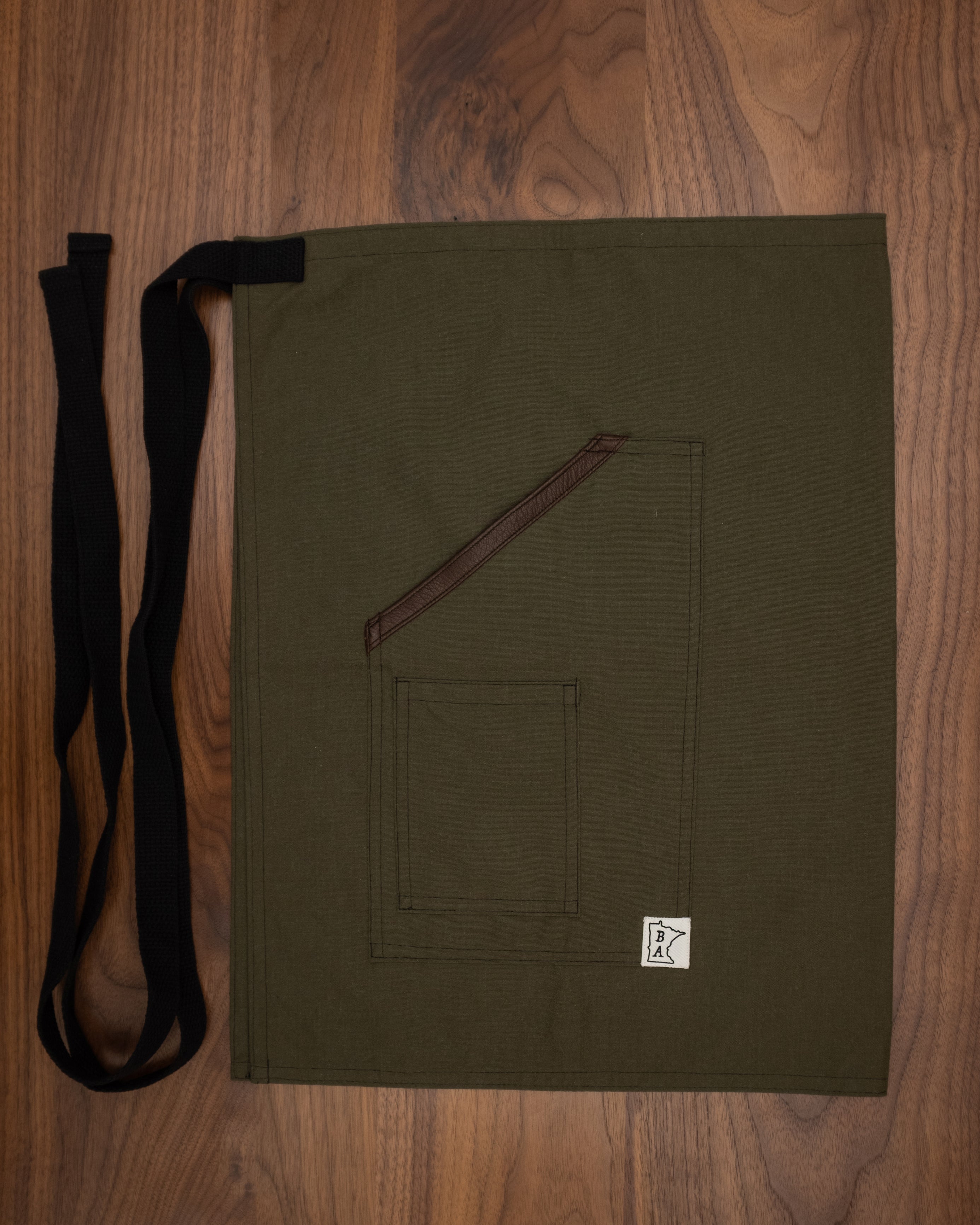 Nomex-Kevlar olive colored bistro apron with black strapping and deerhide trim folded on a wooden surface. The apron design Fire Watch was manufactured in Minnesota by Craftmade Aprons. The apron body protects against flames and punctures.