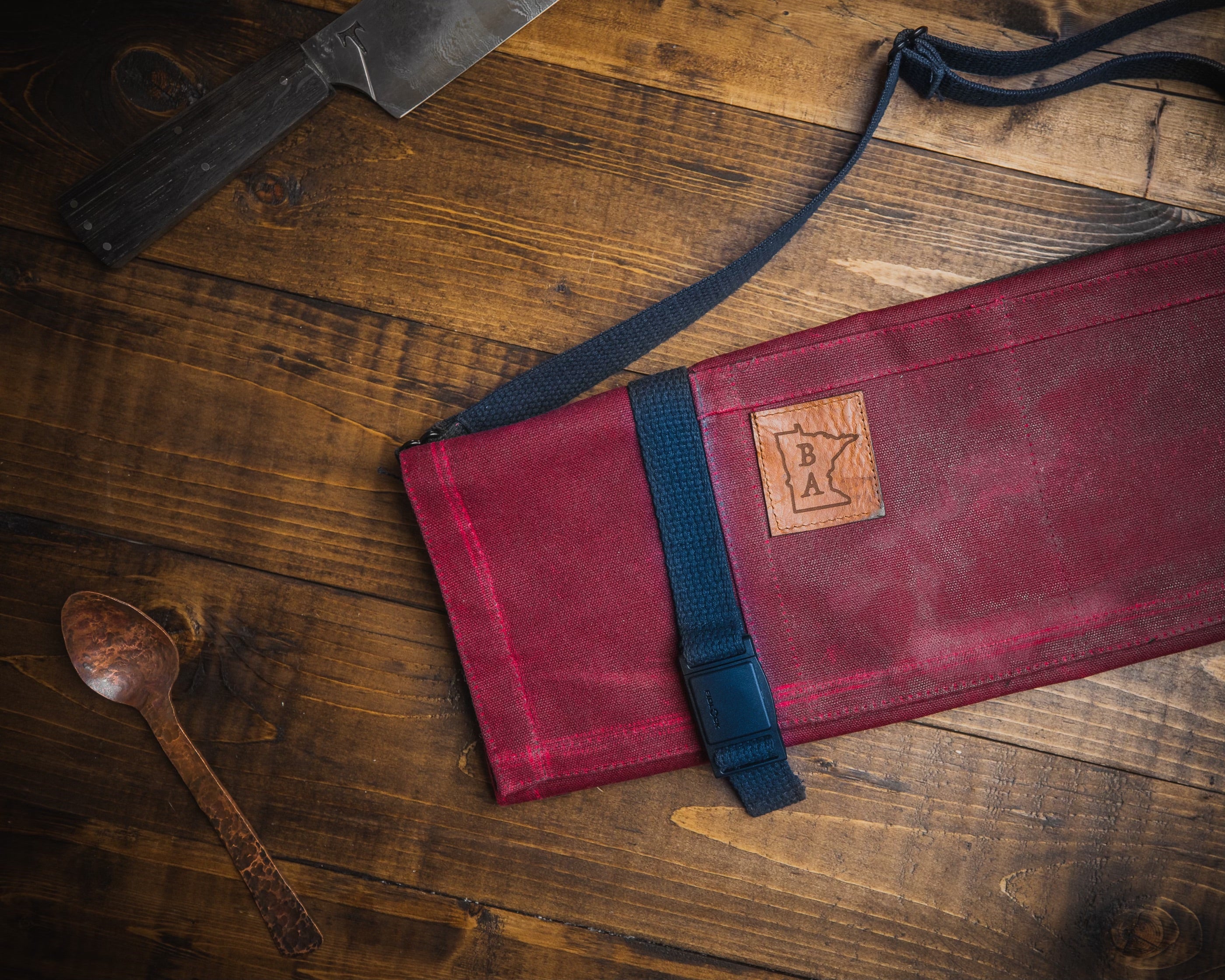 Product shot of the burgundy Main Squeeze roll on a wooden surface next to a knife and copper hammered spoon. The cotton water repellant and flame resistant knife roll was designed by Craftmade Aprons, Minnesota.