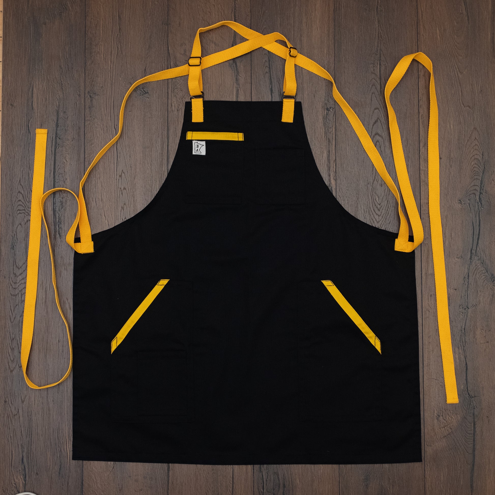 Cotton/poly black apron with yellow trim on the pockets and yellow strapping laid out flat on a wooden surface. Design Killa Beez by Craftmade Aprons, based in Minnesota.