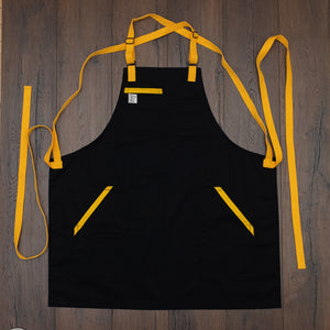 Cotton/poly black apron with yellow trim on the pockets and yellow strapping laid out flat on a wooden surface. Design Killa Beez by Craftmade Aprons, based in Minnesota.