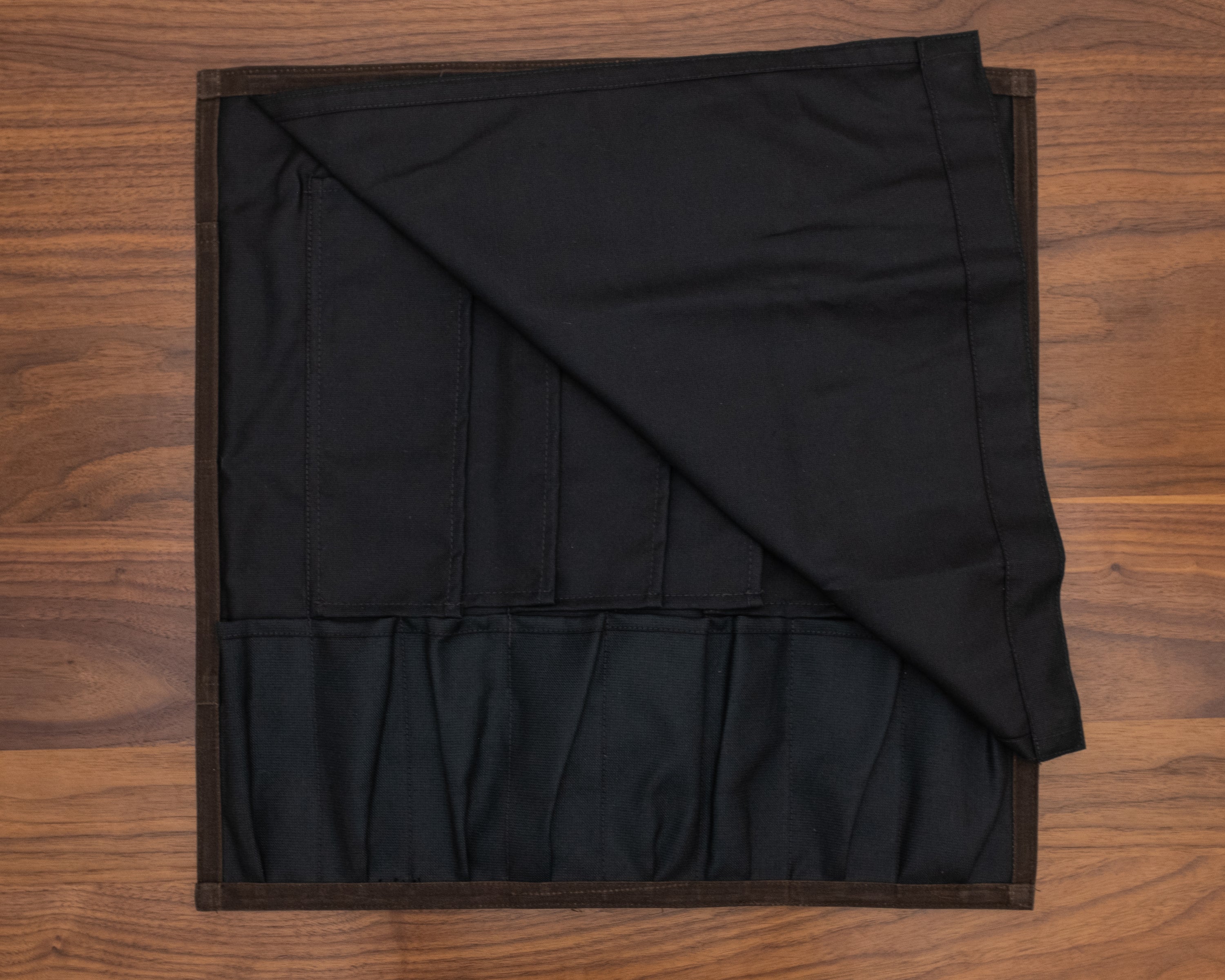 Black interior of the Main Squeeze dark oak knife roll laid out flat on a wooden surface. The interior is made of a 50-50 Kevlar/Nomex blend for long lasting protection. Design by Craftmade Aprons in Minnesota.