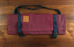 Cotton burgundy knife roll protected by tex-wax with navy blue strapping displayed on a wooden surface. The Main Squeeze knife roll was manufactured in Minnesota by Craftmade Aprons. 