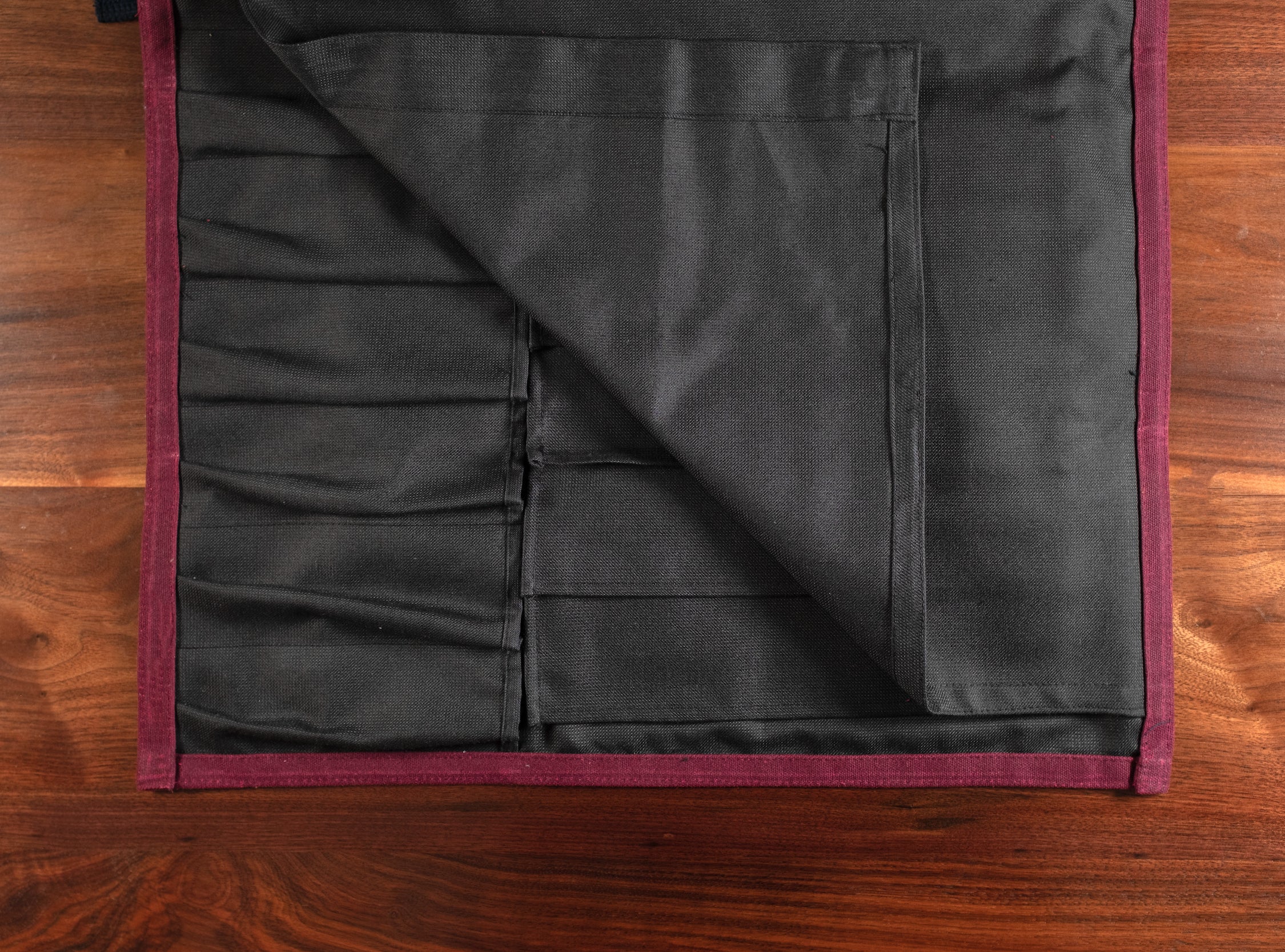 The interior of the Main Squeeze burgundy knife roll laid out flat on a wooden surface. The interior is a 50-50 Kevlar/Nomex blend. Every stitch made by Craftmade Aprons, Minnesota.