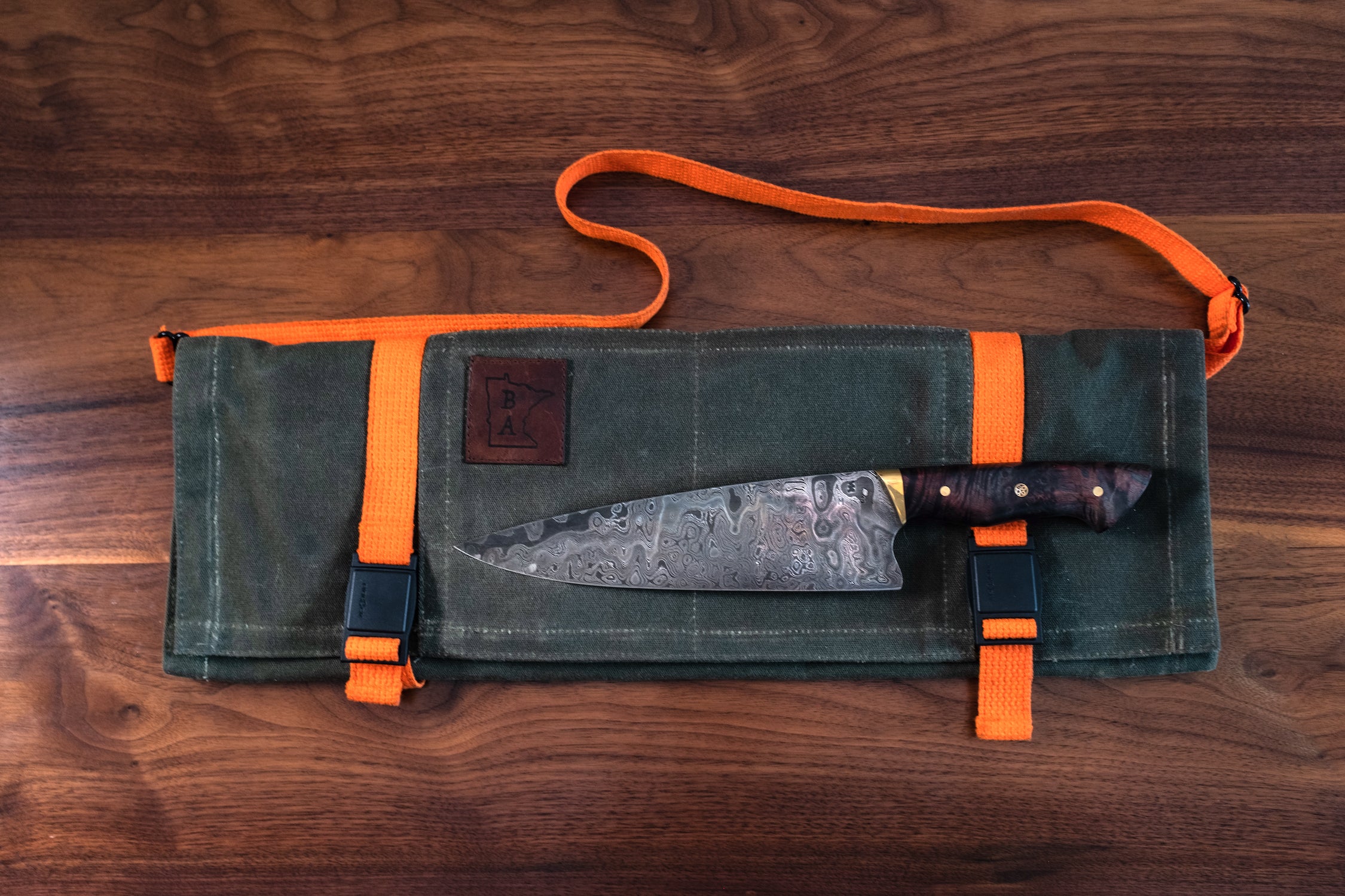 Knife laying on the exterior of the olive green tex-wax protected Main Squeeze knife roll laying on a wooden surface. Manufactured in Minnesota by Craftmade Aprons. 