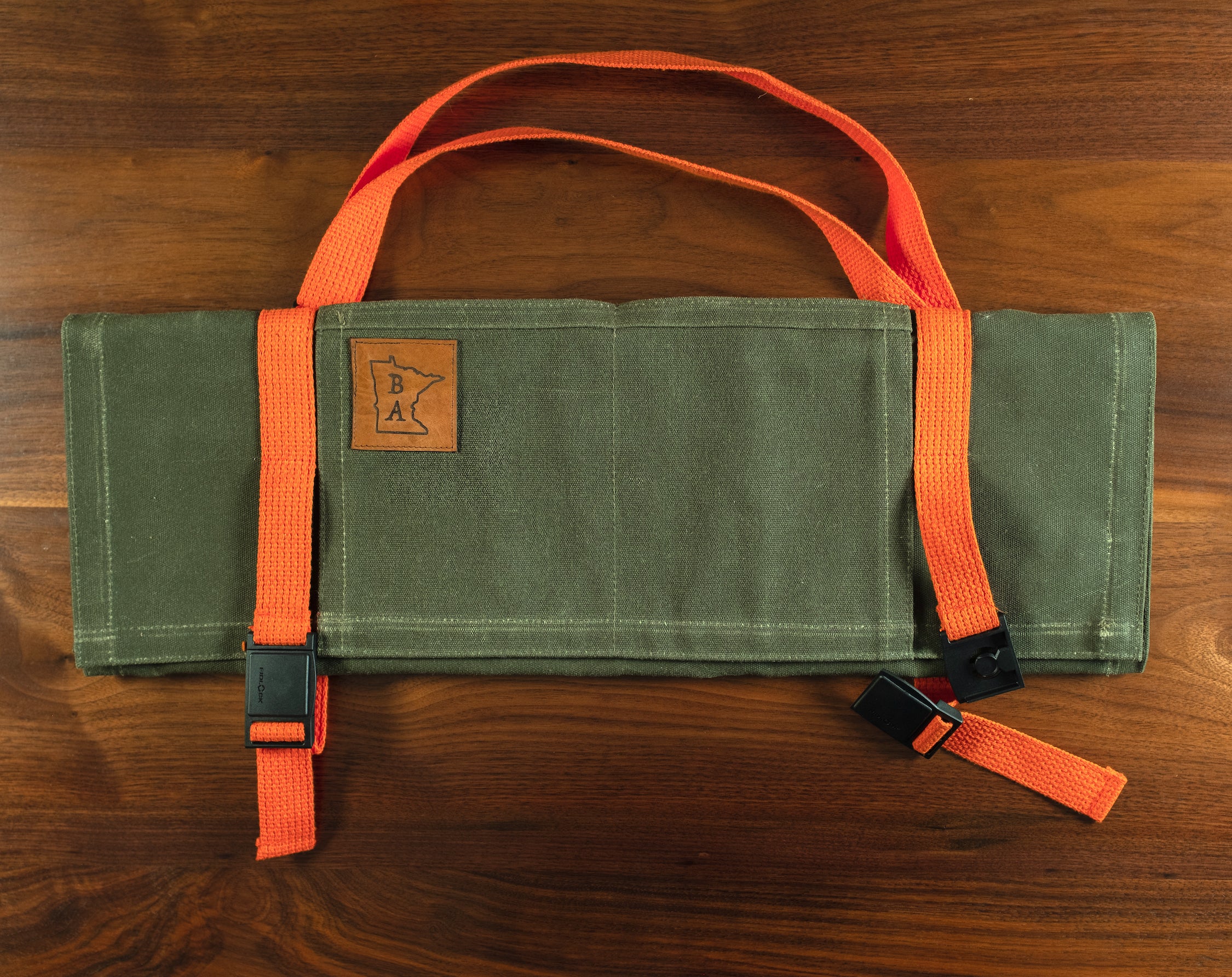 The Main Squeeze olive green cotton knife roll folded on a wooden surface with the buckles released. The knife roll was designed and produced by Craftmade Aprons in Minnesota.
