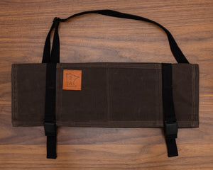 Cotton dark oak knife roll protected in tex-wax folded with the black strapping on a wooden surface. The knife roll design Main Squeeze was manufactured in Minnesota by Craftmade Aprons.