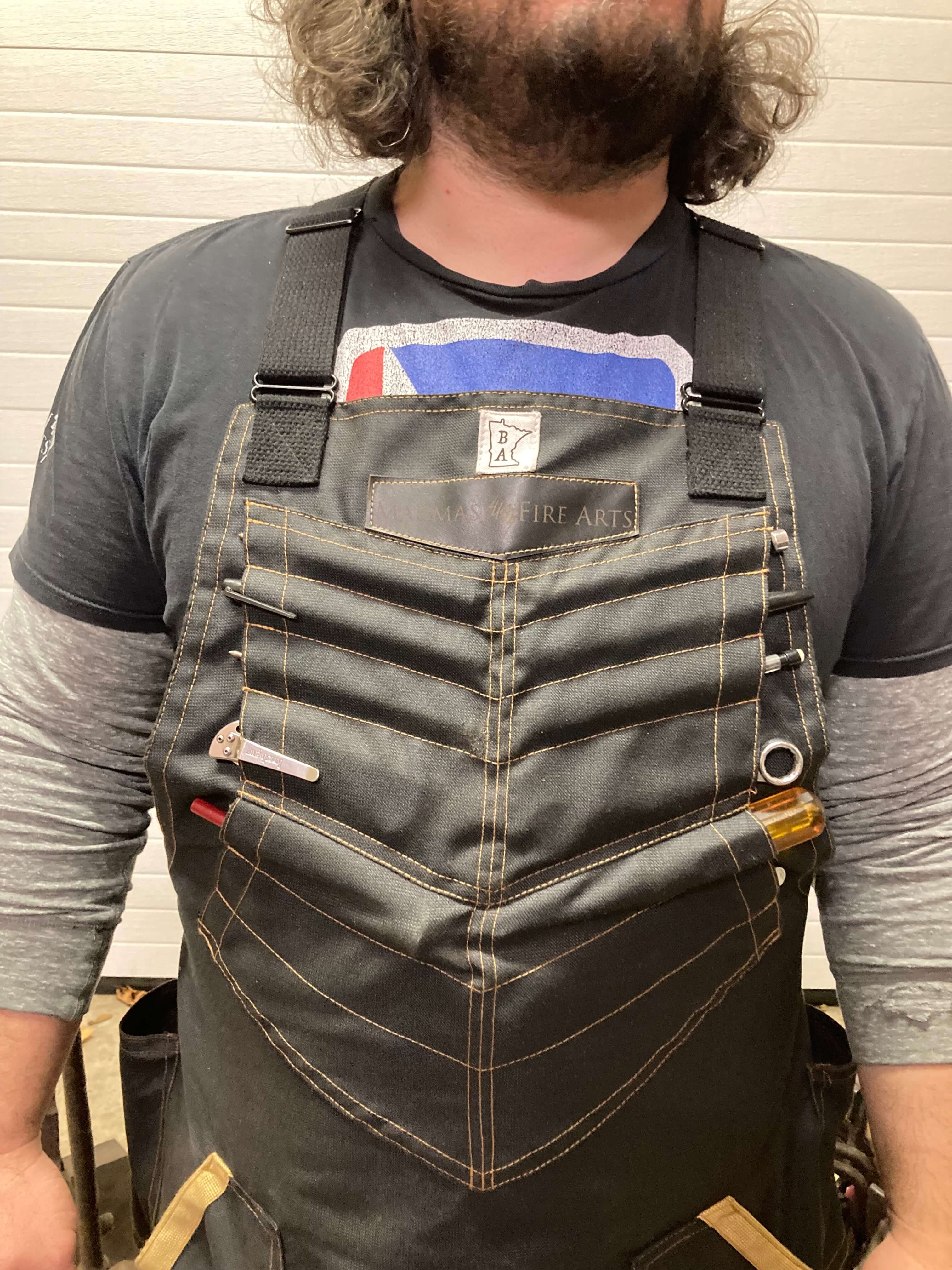 Image of Mareko Maumasi modelling the BAMF apron, with emphasis on the apron's upper tool pockets. The BAMF is a heavy-duty, protective Kevlar apron in black with yellow threading, and the perfect protective apron for knife makers.