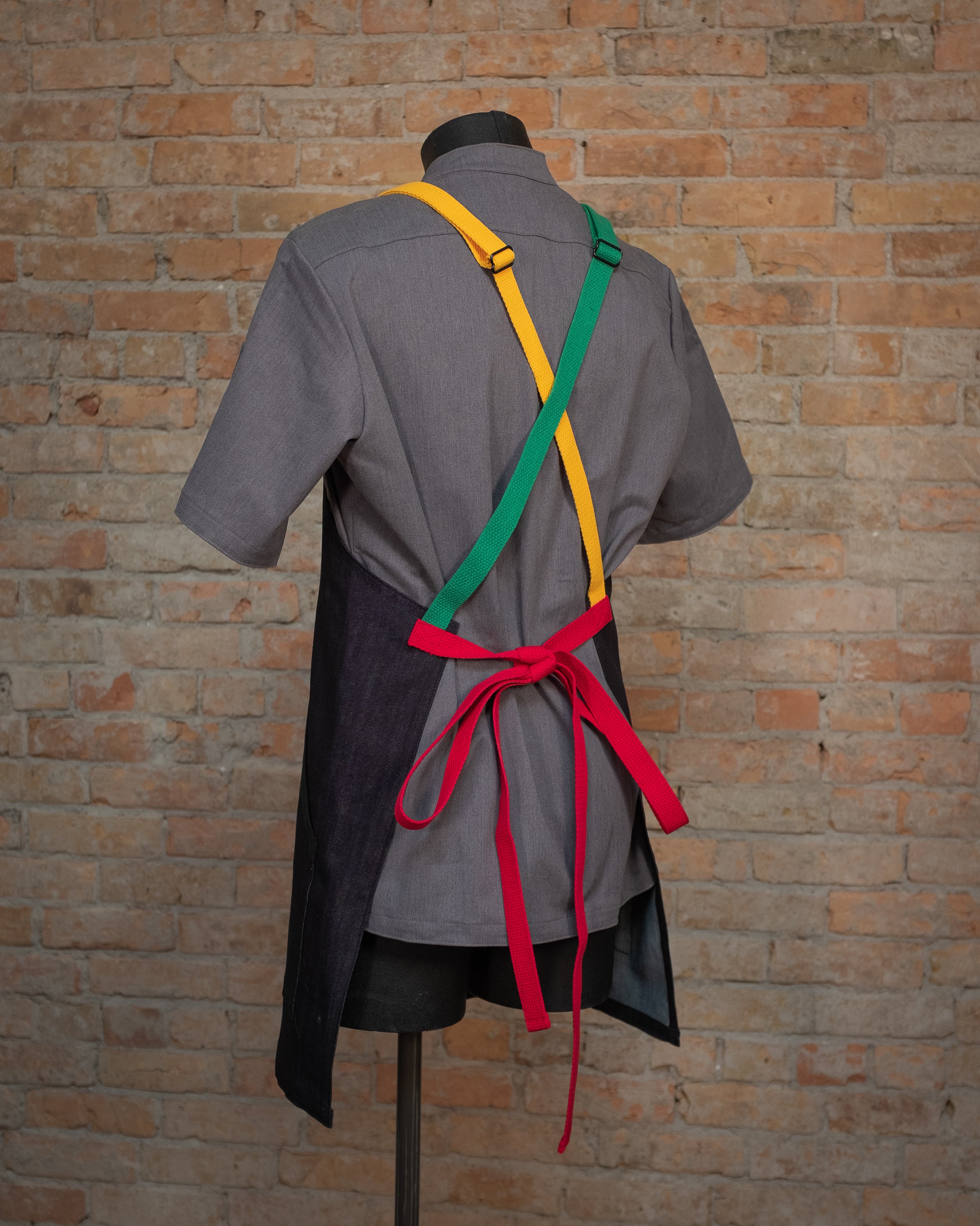 Denim crossback apron from Craftmade Aprons, Minnesota, displayed on a mannequin over a gray chef coat. The apron Marley is shown from the back, accentuating the red, green, and yellow trio crossback strapping.