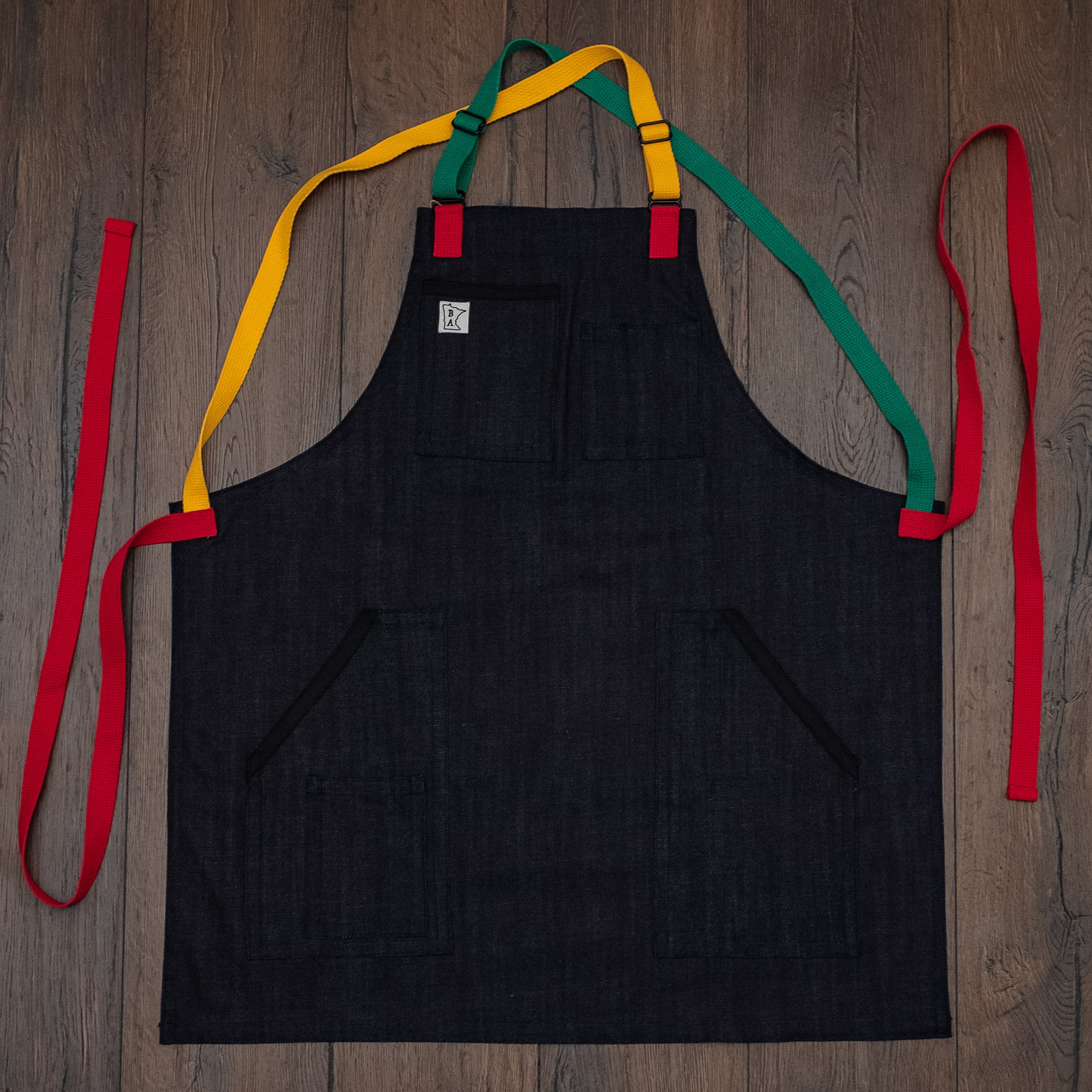 Denim apron with black trimmed pockets and red, green, and yellow strapping laid out flat on a wooden surface. Design Marley by Craftmade Aprons, based in Minnesota.