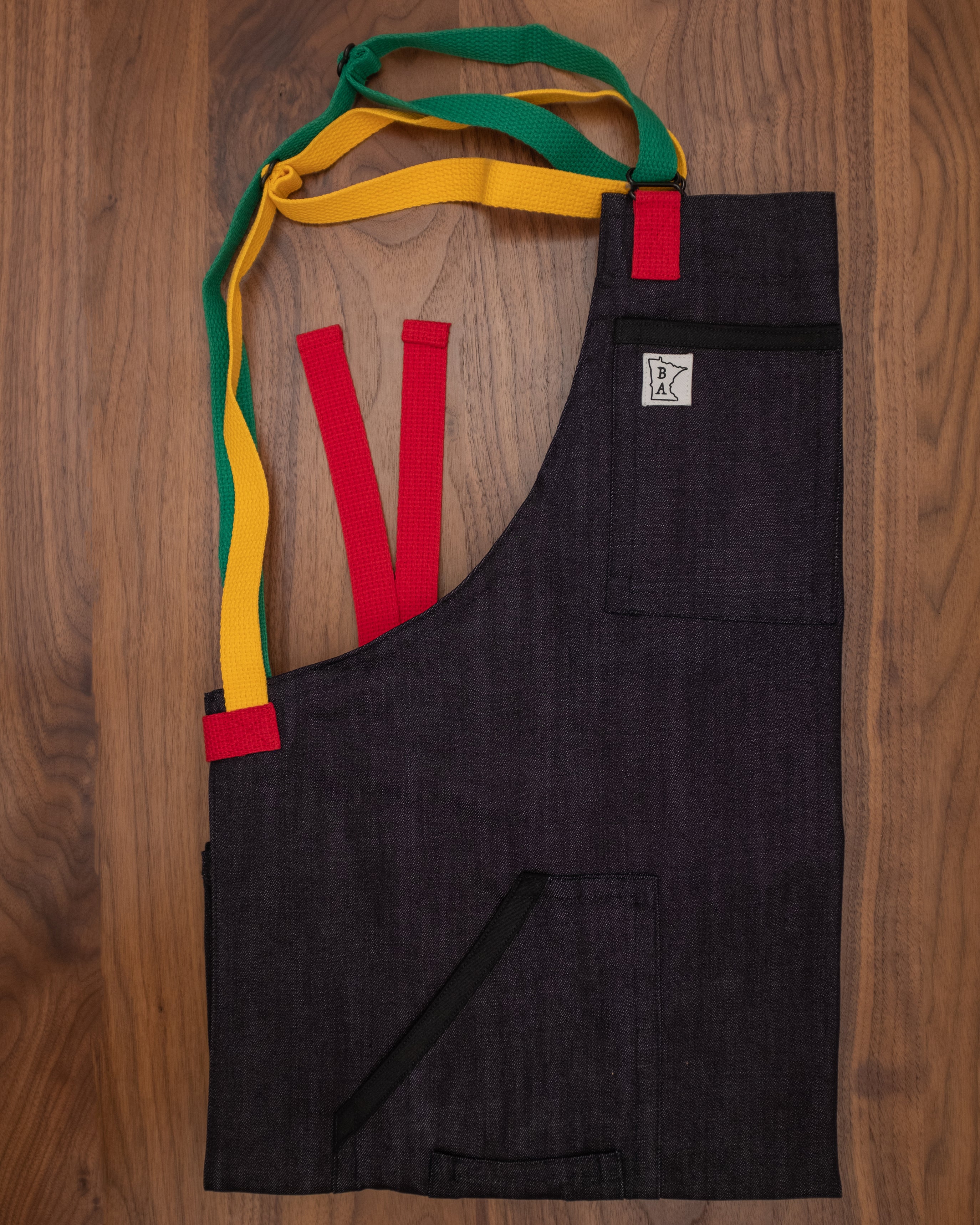 Denim apron with black trimming on the pockets and a trio of red, yellow, and green strapping folded on a wooden surface. The apron design Marley was manufactured in Minnesota by Craftmade Aprons. 