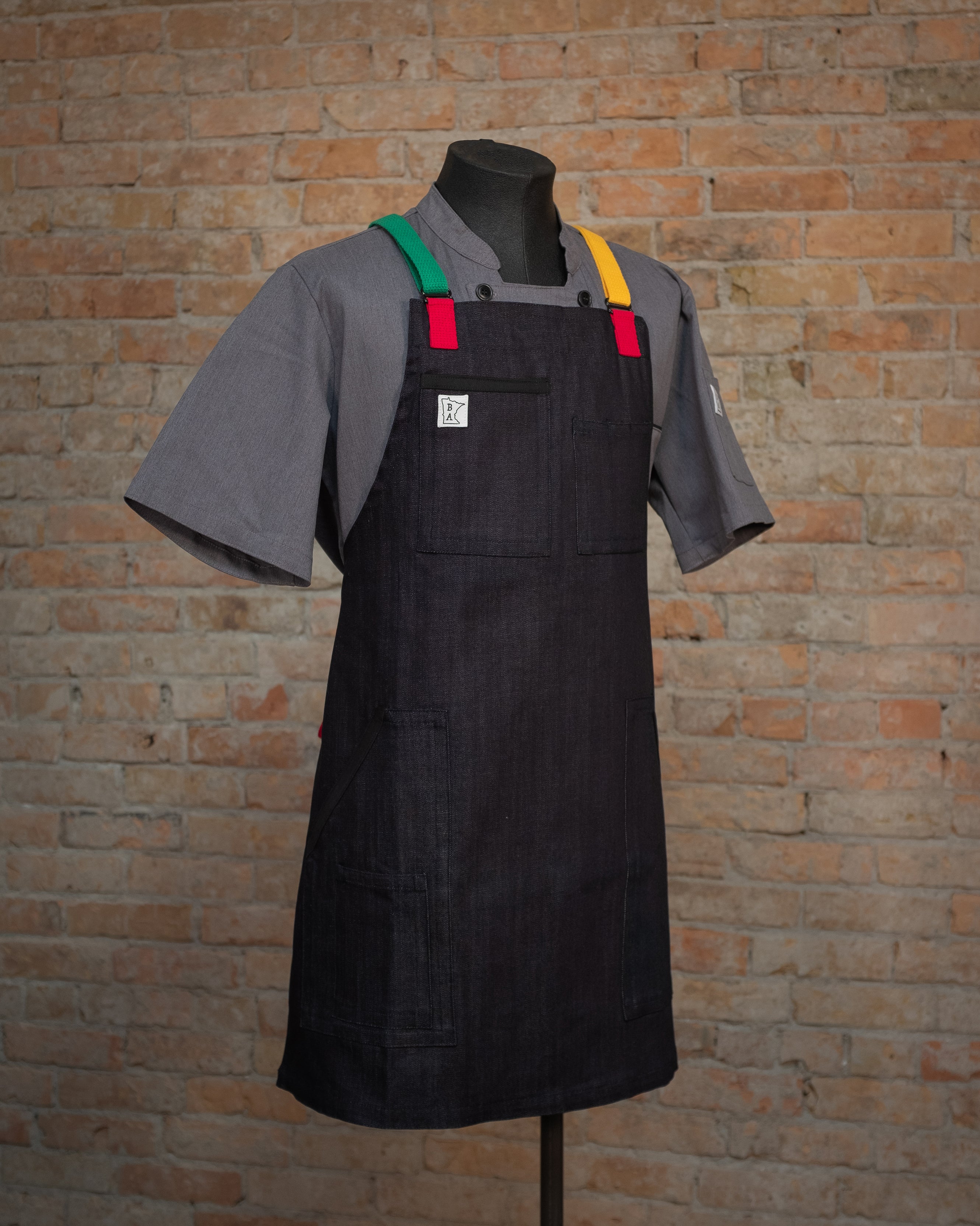 The denim apron design Marley displayed on a mannequin over a gray chef coat. Both the chef coat and the apron were designed and produced by Craftmade Aprons, Minnesota. 