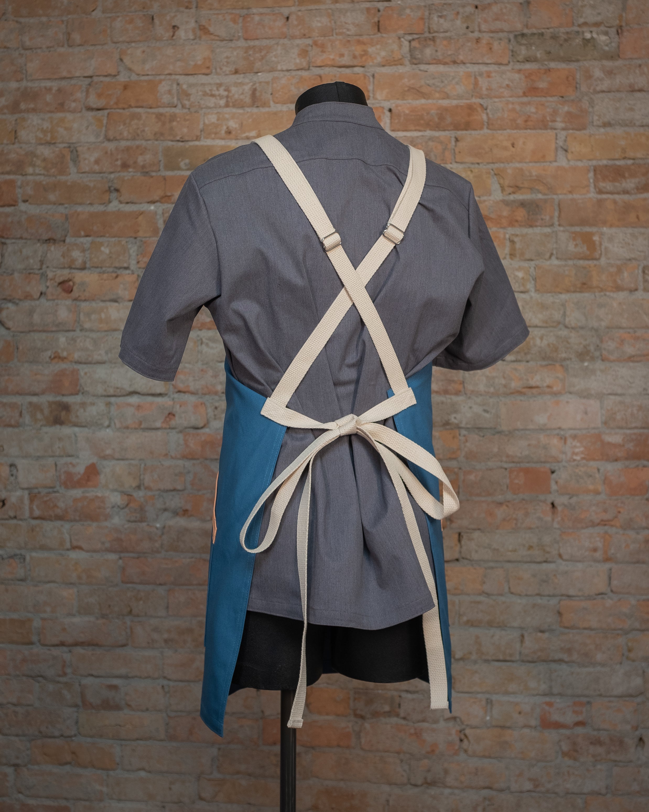 Cotton duck cloth apron from Craftmade Aprons, Minnesota displayed on a mannequin over a gray chef coat. The apron Miami Nice is shown from the back, displaying the tan crossback strapping.