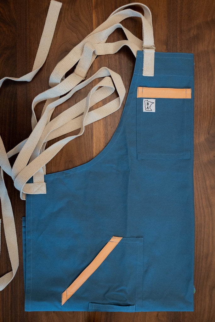 Blue cotton duck cloth apron with peachy orange trim and tan strapping folded on a wooden surface. The apron design Miami Nice was manufactured in Minnesota by Craftmade Aprons.