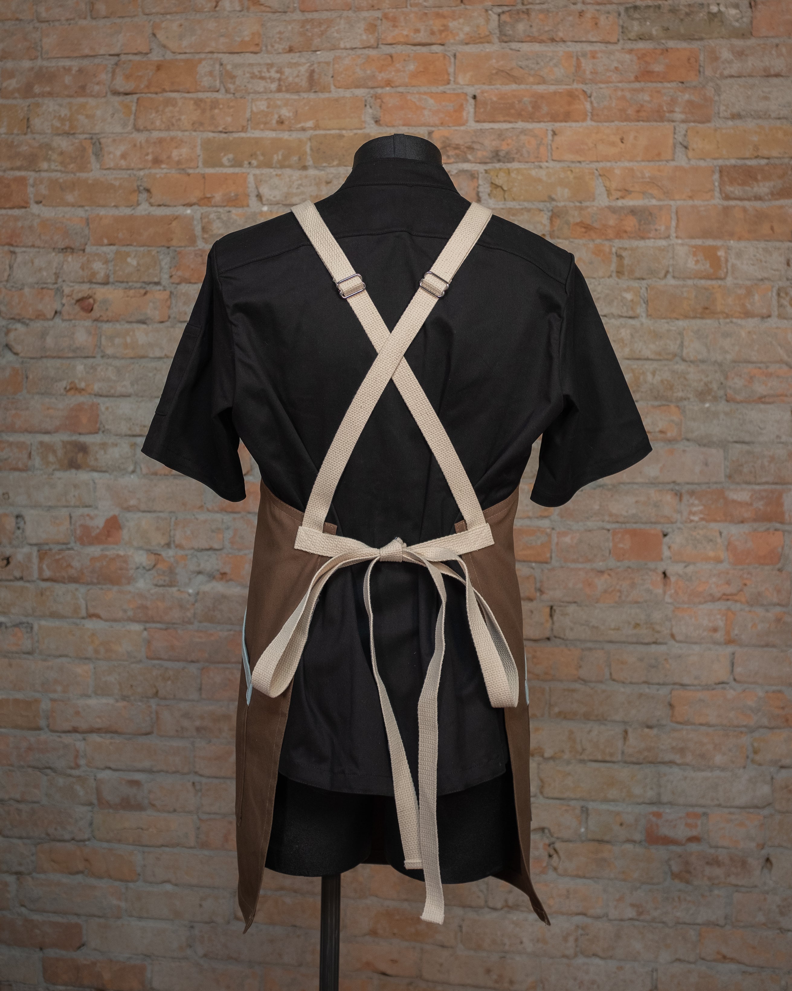 Duck duck cloth crossback apron from Craftmade Aprons, Minnesota, displayed on a mannequin over a black chef coat. The apron Sea Mist is shown from the back, displaying the natural cotton crossback strapping.