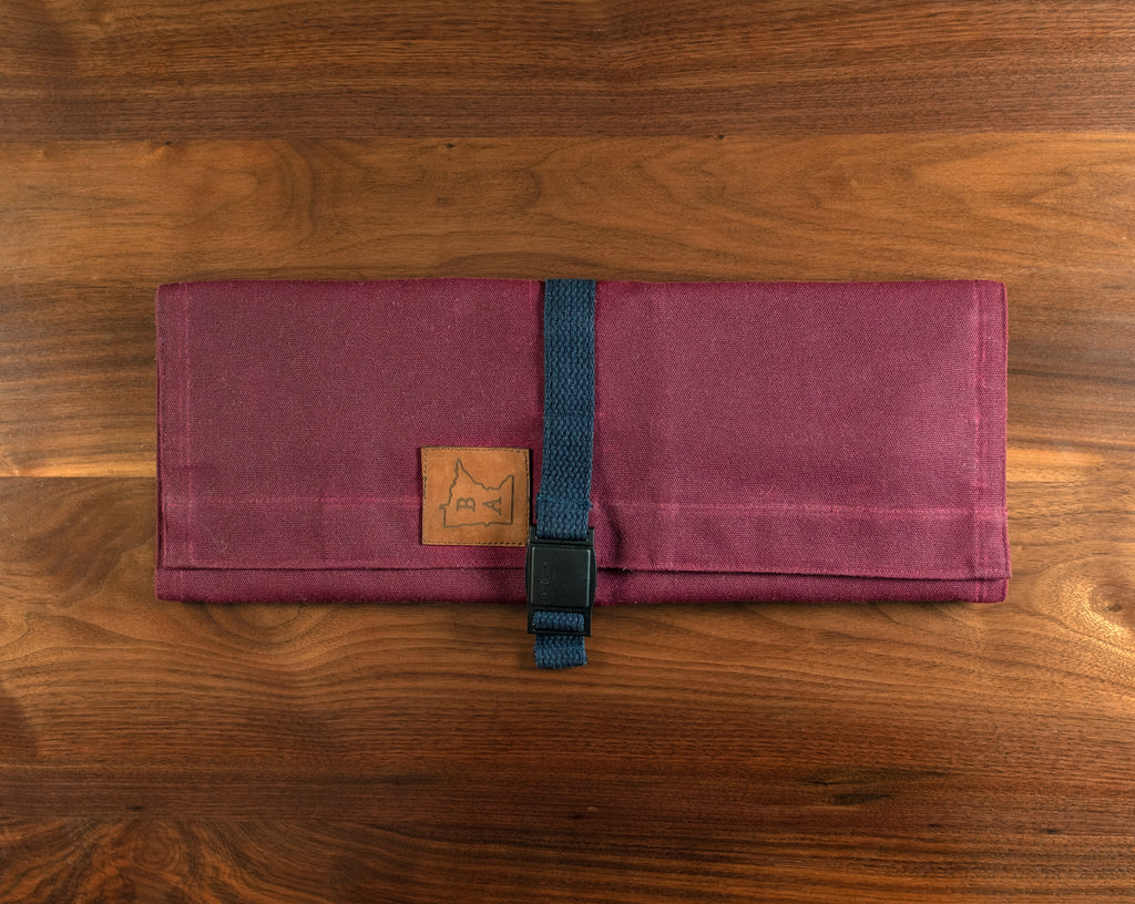 Cotton burgundy knife roll protected by tex-wax with navy blue strapping displayed on a wooden surface. The Side Hustle knife roll was manufactured in Minnesota by Craftmade Aprons. 