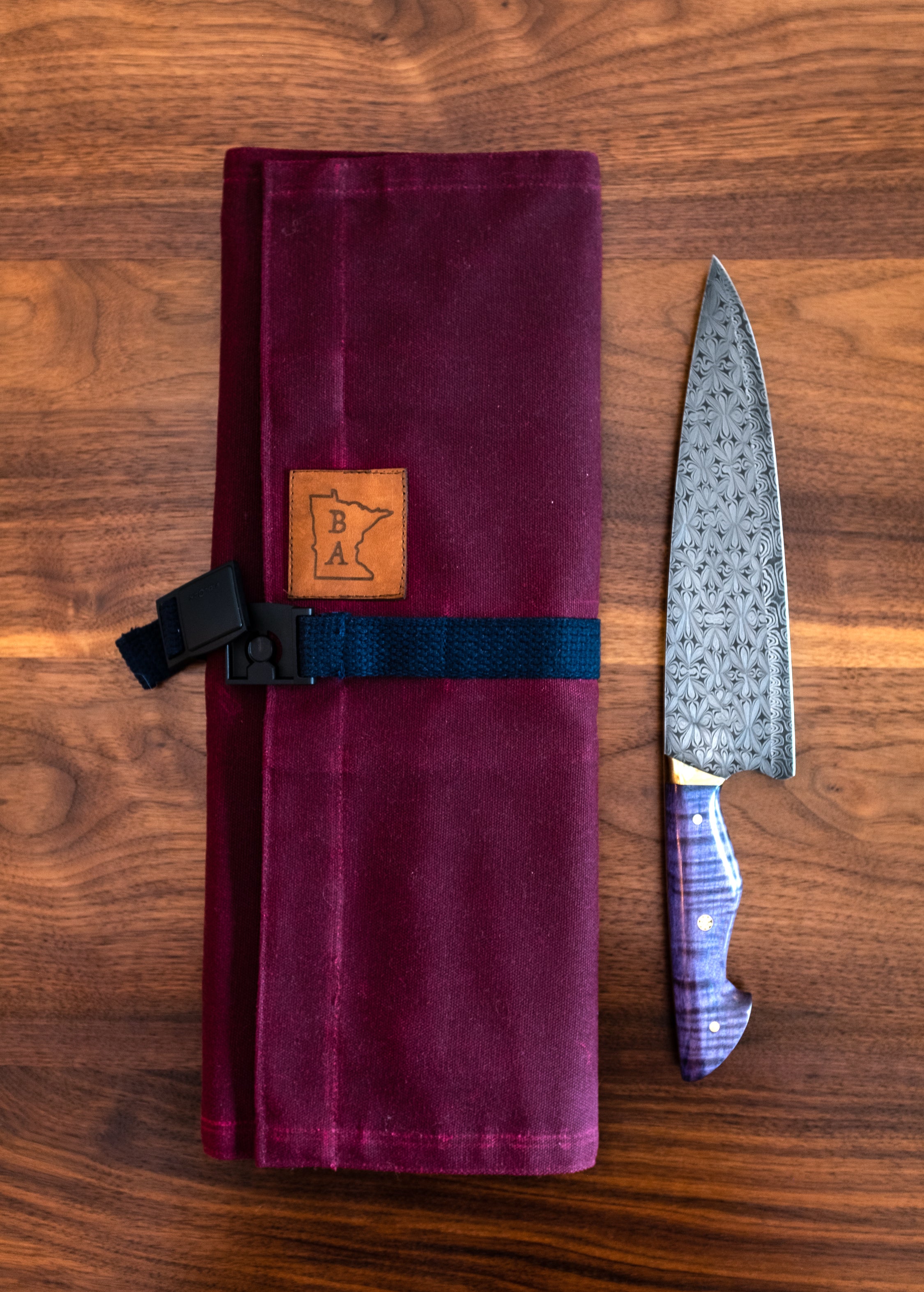 Product shot of burgundy Side Hustle knife roll buckled on a wooden surface next to knife. Handcrafted in Minnesota by Craftmade Aprons.